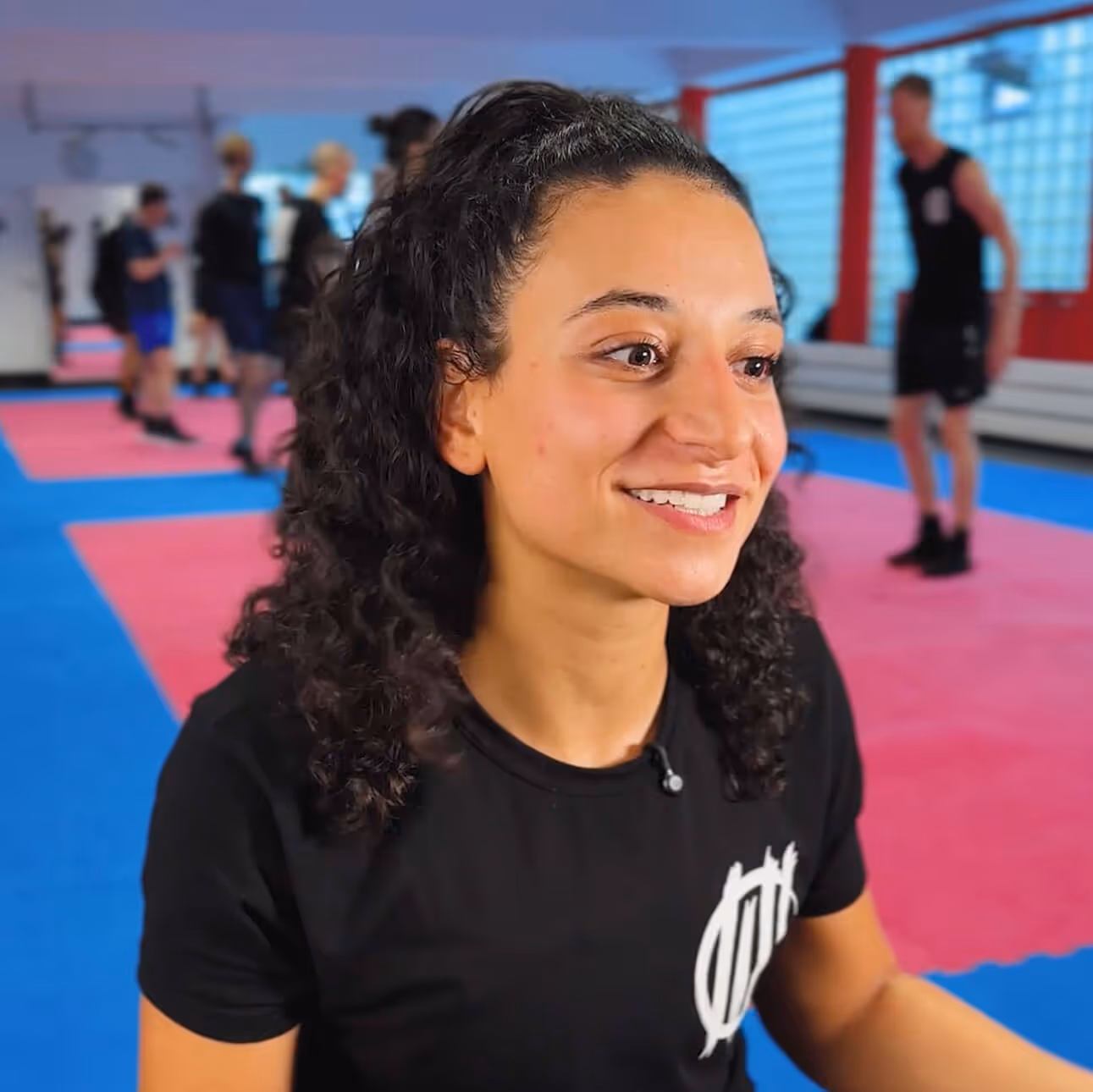 Lächendes Mädchen mit braunem T-Shirt sitzt auf blauroter Matte in einem Kampfsportstudio.