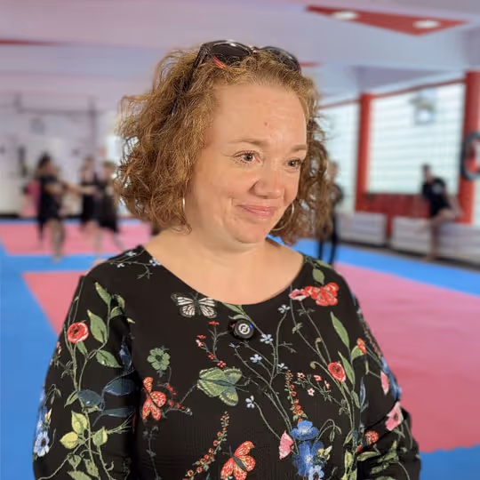 Frau mit lockigem Haar trägt eine schwarze Bluse mit bunten Blumen- und Schmetterlingsmustern in einem Raum mit blau-rotem Boden.