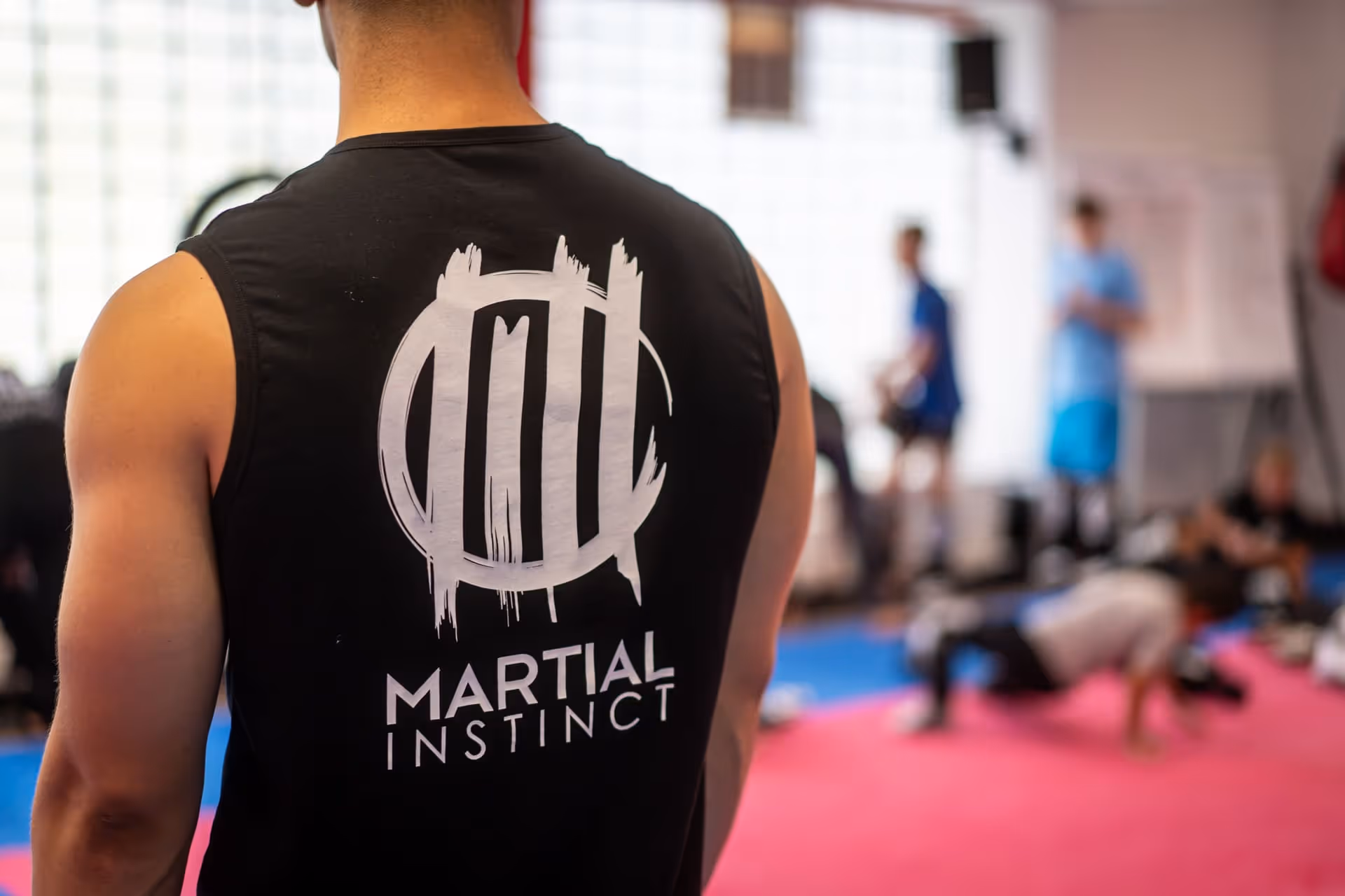 Rückansicht eines Mannes mit einem schwarzen ärmellosen Shirt mit dem Schriftzug 'Martial Instinct' in einem Trainingsraum mit anderen Personen im Hintergrund.