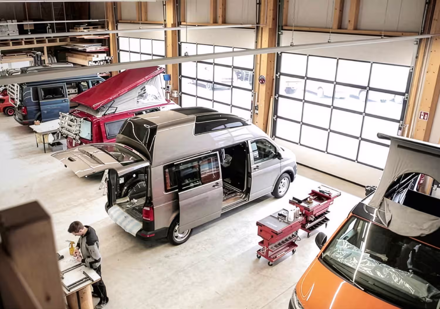 Werkstatt mit mehreren Campingbussen, darunter ein grauer und ein orangefarbener mit geöffnetem Aufstelldach, und einem Mann bei der Arbeit.