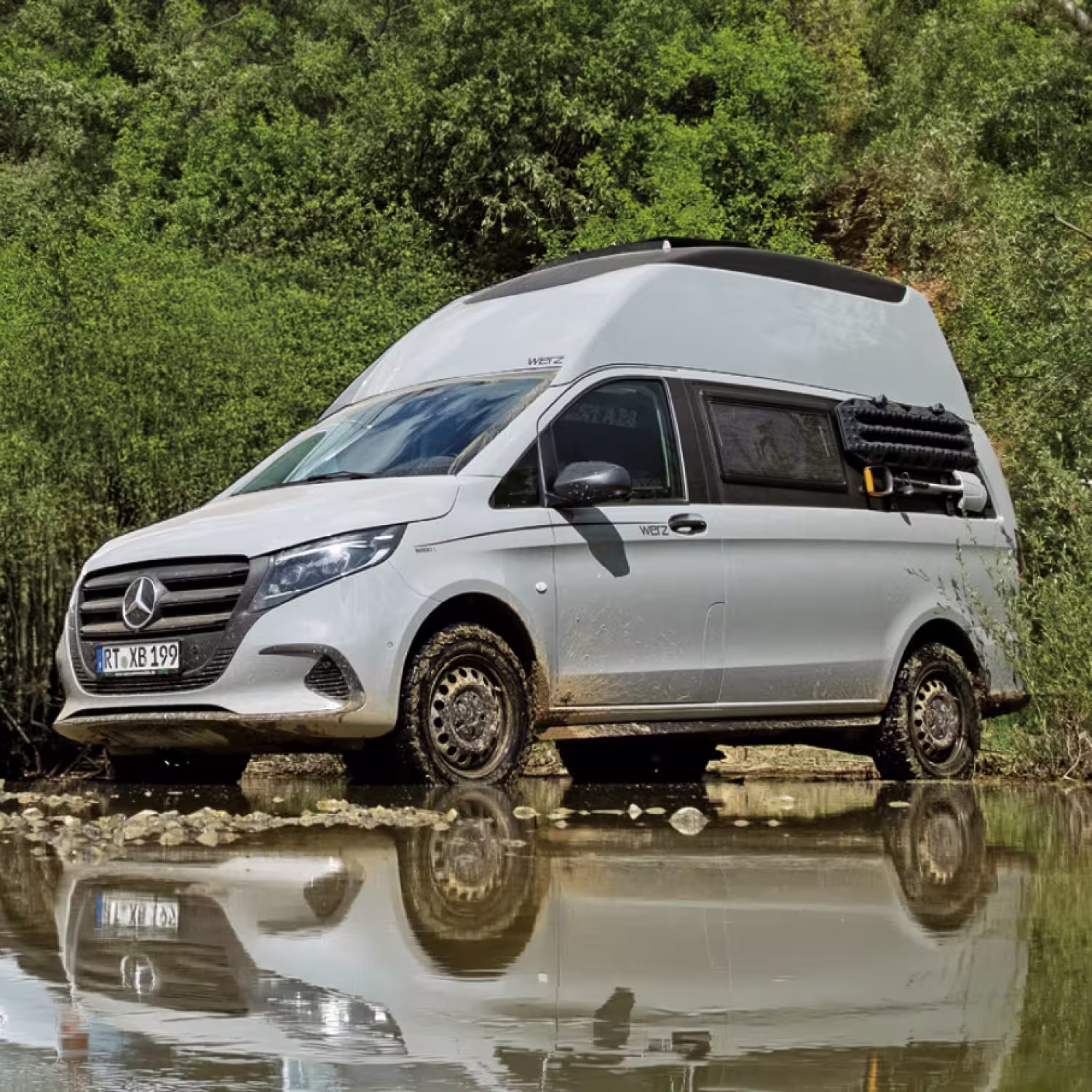 Weißer Mercedes-Camper mit hohem Dach und verschmutzten Reifen steht am Ufer eines flachen Gewässers mit Spiegelung im Wasser.