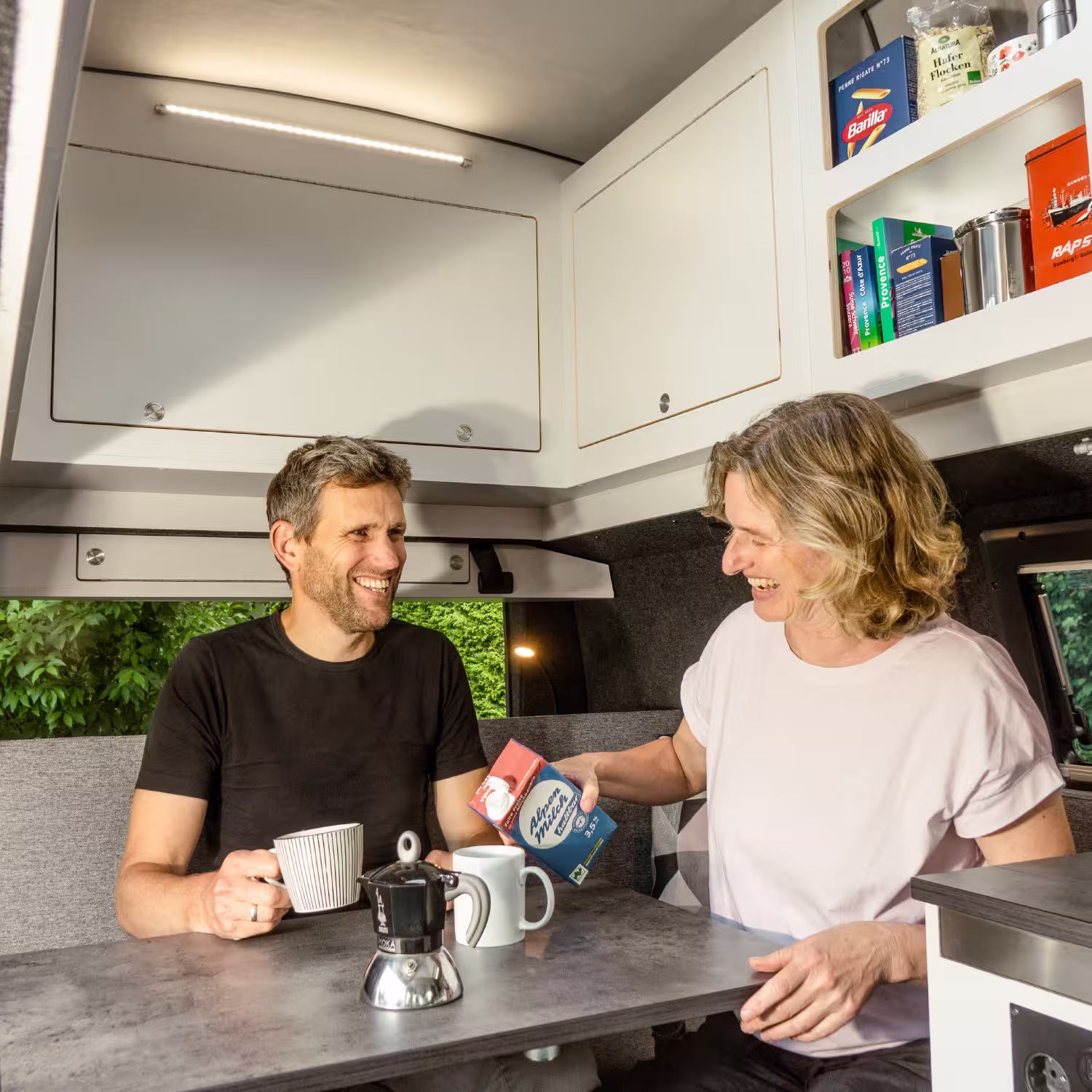 Paar lacht beim Kaffeetrinken im Wohnmobil, sie hält eine Milchpackung, er eine Tasse.