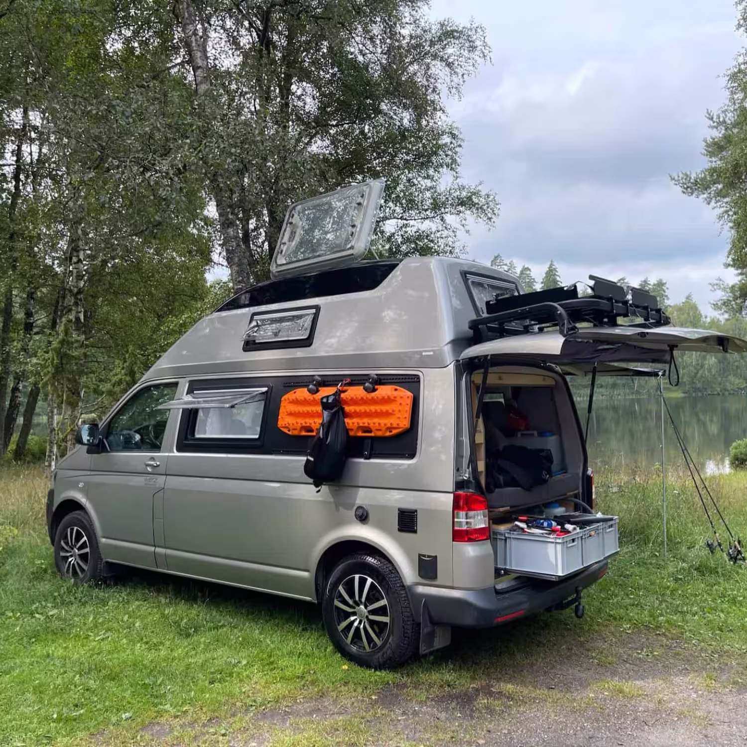 Silberner Camper-Van mit geöffnetem Dachfenster und Heckklappe, geparkt auf einer Wiese neben einem See mit Werkzeugkiste im Kofferraum.