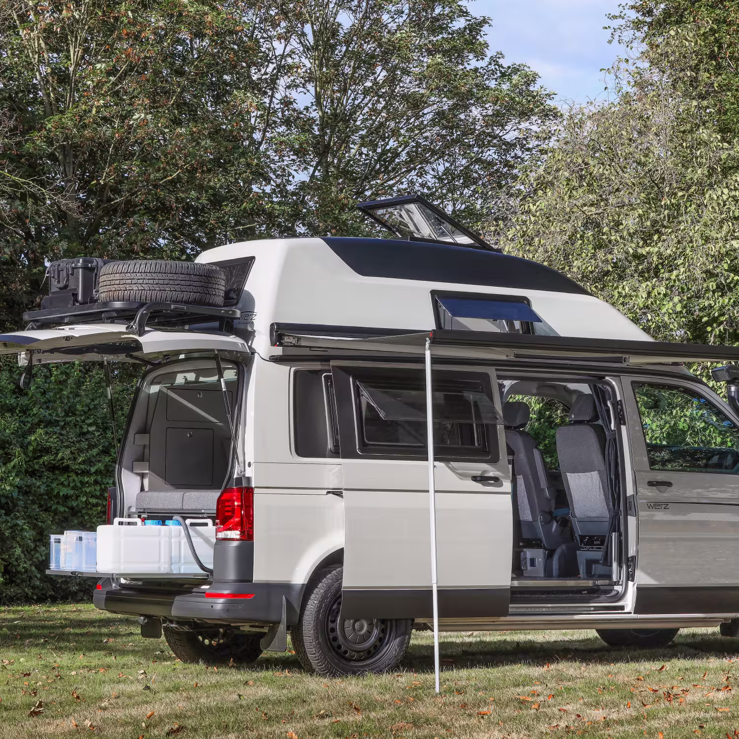 Wohnmobil mit geöffneten Türen, ausgefahrenem Sonnenschutz und aufgeklapptem Dachfenster auf einer Wiese vor Bäumen.