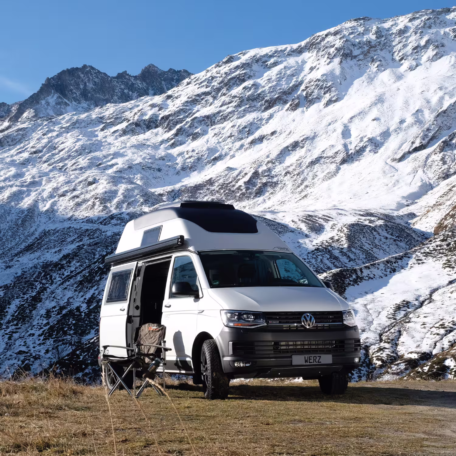 Weißer Volkswagen Campervan mit geöffnetem Seitentür und Klappstuhl davor, stehend auf grasigem Platz vor verschneiten Bergen.