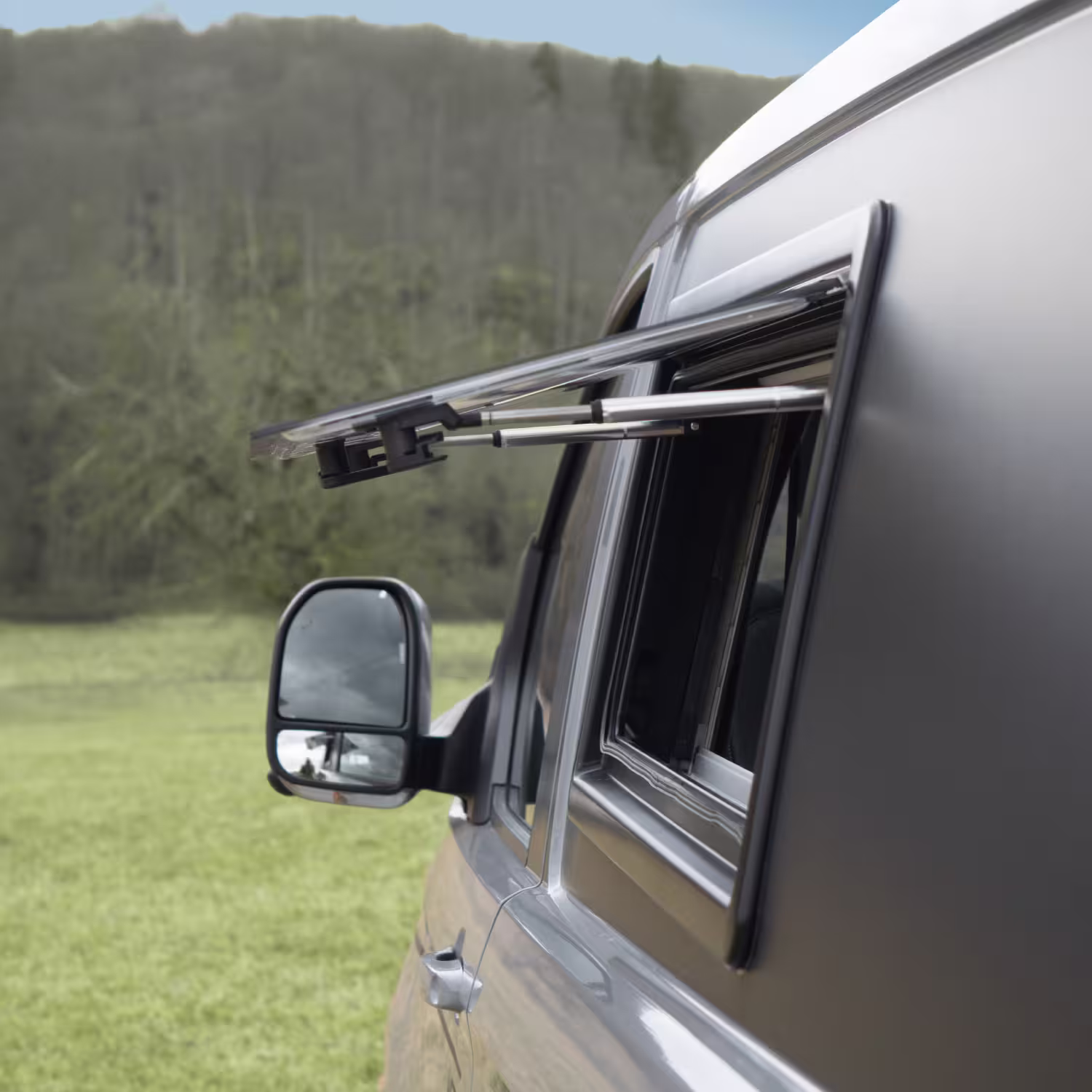 Offenes Fenster mit ausklappbarem Regenschutz an einem grauen Camper, im Hintergrund ein grünes Feld und Wald.