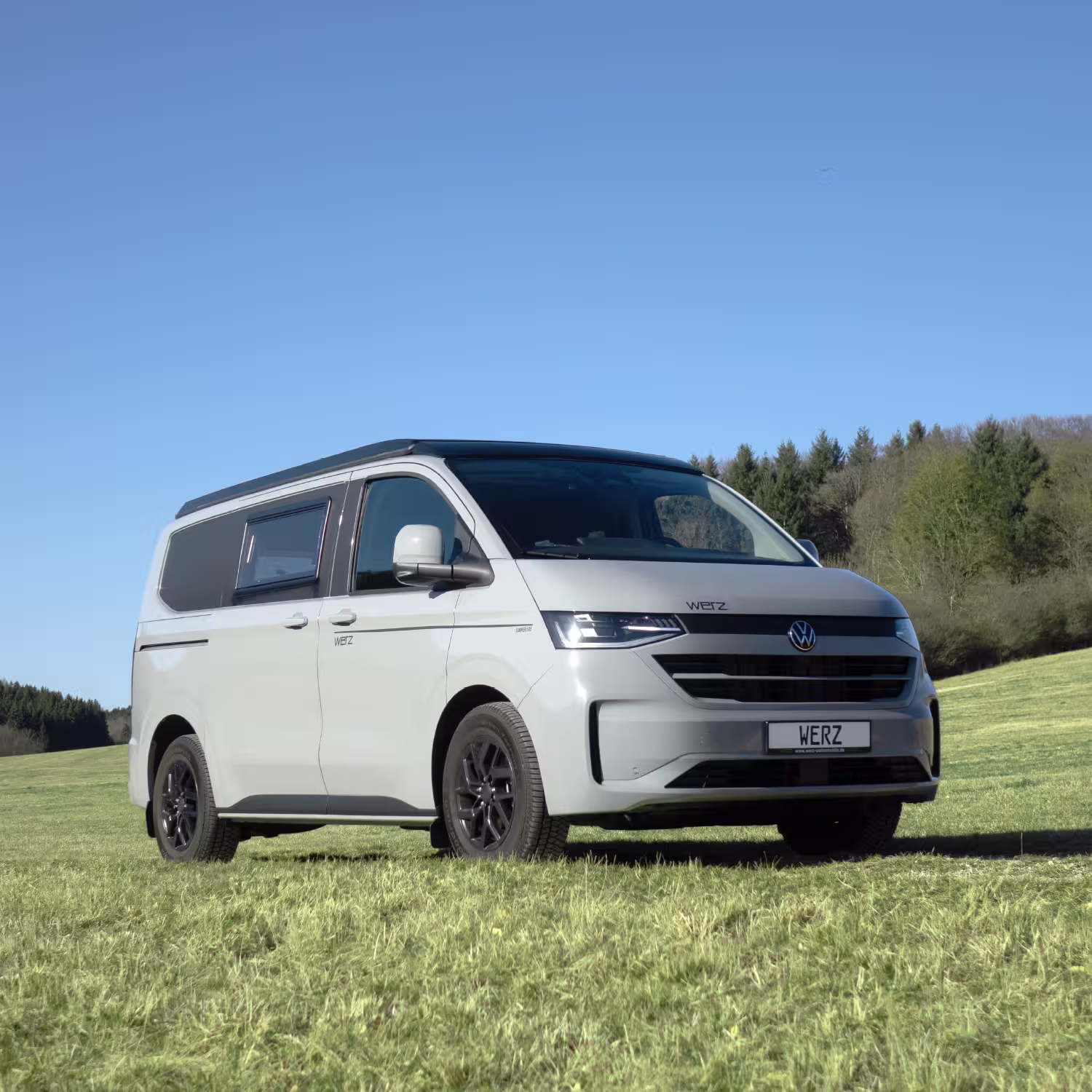 Moderner weißer Werz Camper-Van steht auf einer grünen Wiese mit Wald und blauem Himmel im Hintergrund.