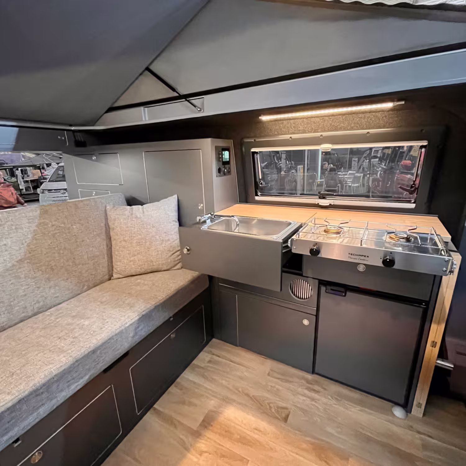 Kompakte Wohnmobilküche mit Spüle, zweiflammigem Gasherd, Kühlschrank und Sitzbank mit Kissen unter einem Fenster.