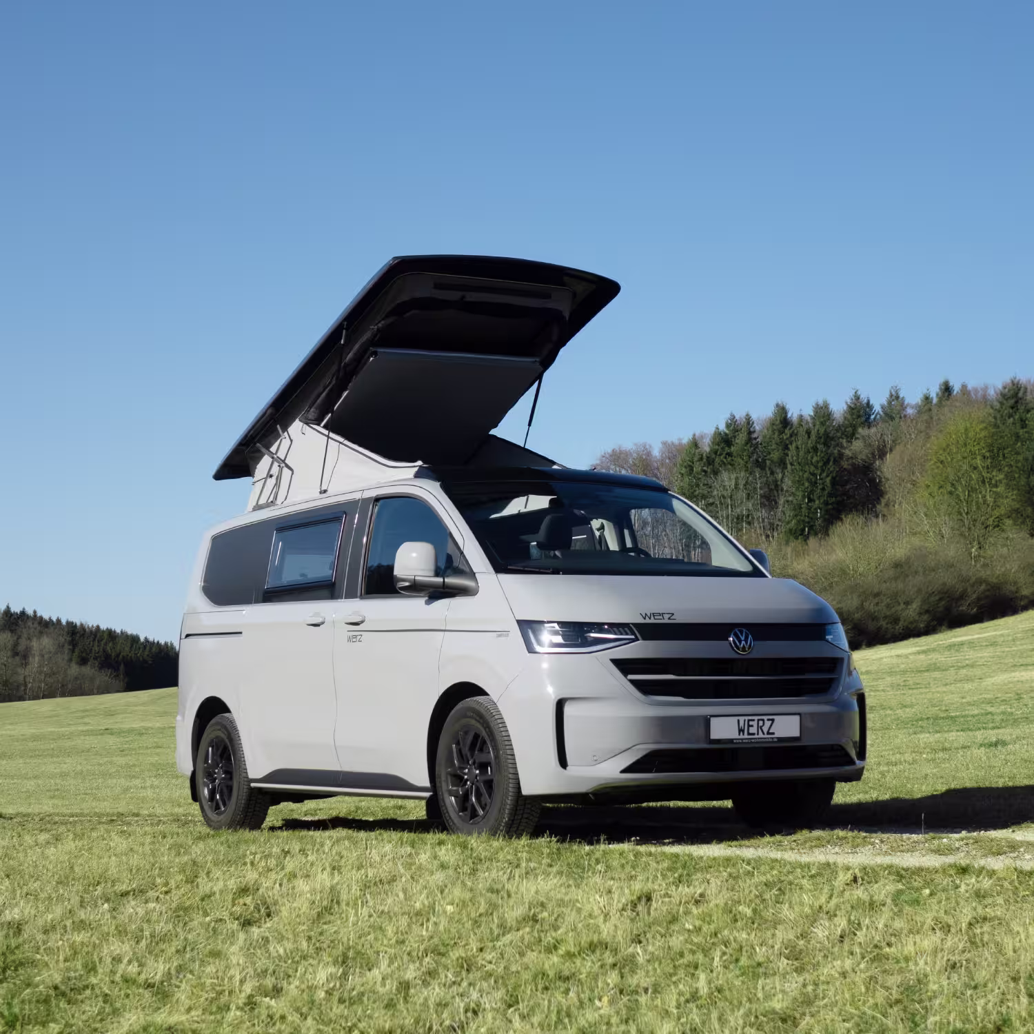 Grauer WERZ Campingbus mit aufgeklapptem Aufstelldach auf einer grünen Wiese unter blauem Himmel.