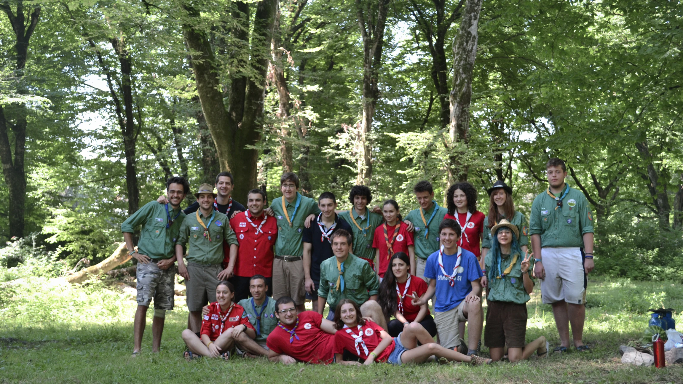 Gruppo di scout sorridenti con divise rosse, verdi e blu in una radura alberata.