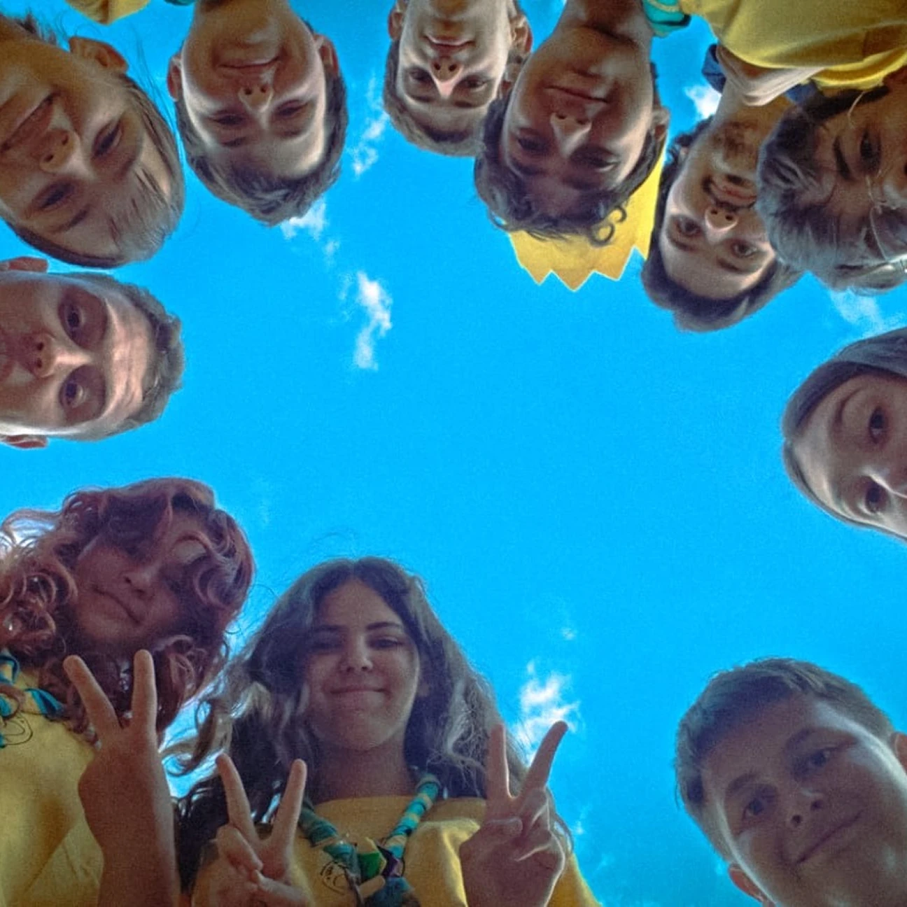 Gruppo di giovani scout in magliette gialle guardano verso la fotocamera con il cielo blu sullo sfondo, alcuni fanno il segno di pace con le dita.