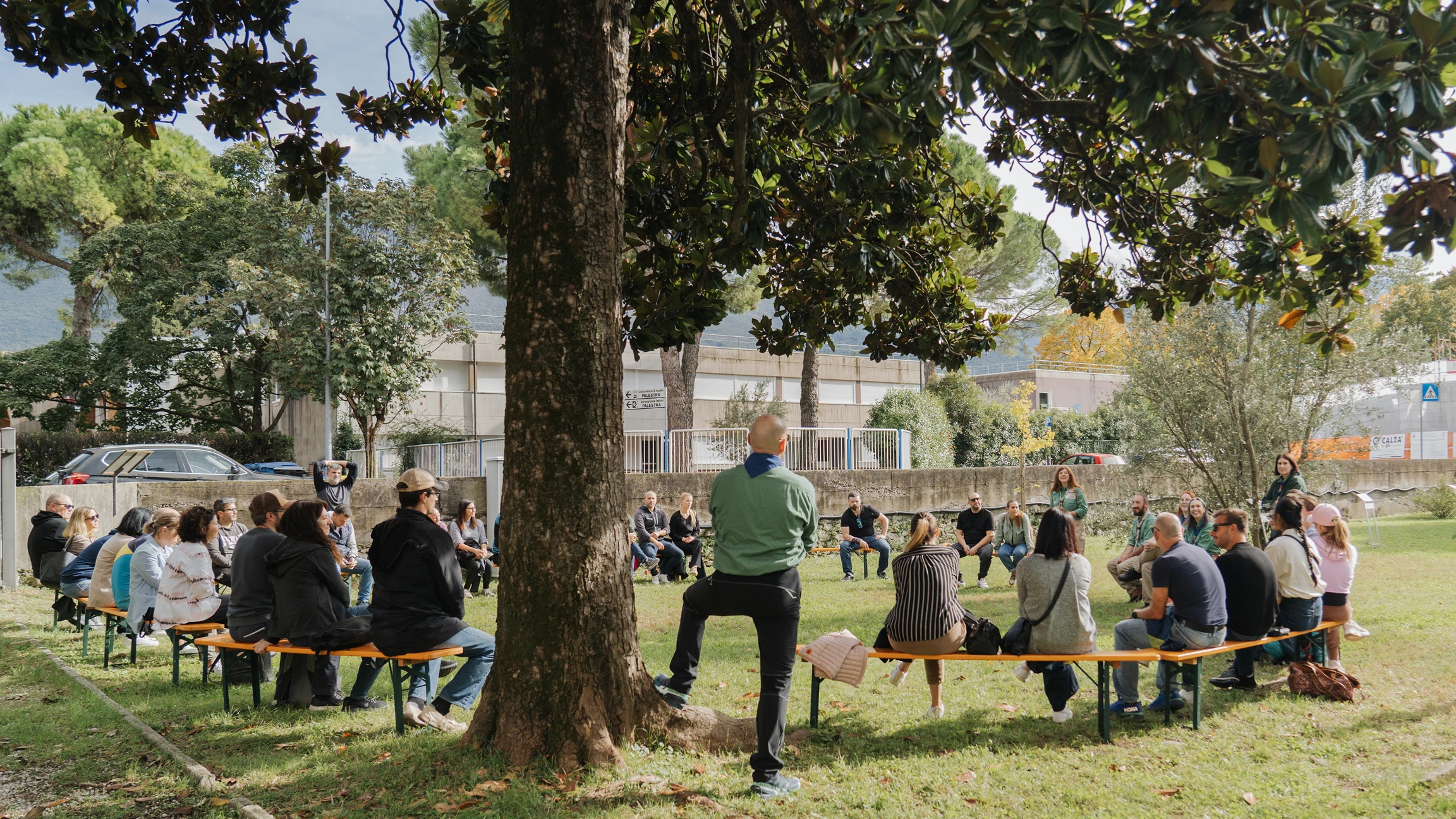 Persone sedute in cerchio su panchine arancioni in un parco all'aperto sotto un grande albero.