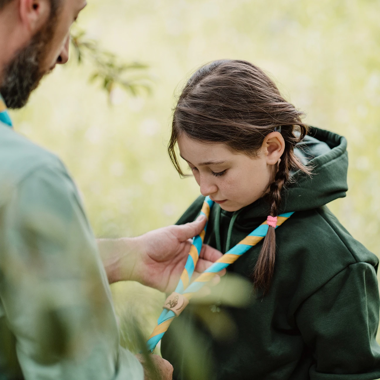Un adulto allaccia un fazzoletto scout blu e giallo al collo di una ragazza con una felpa verde e capelli intrecciati.