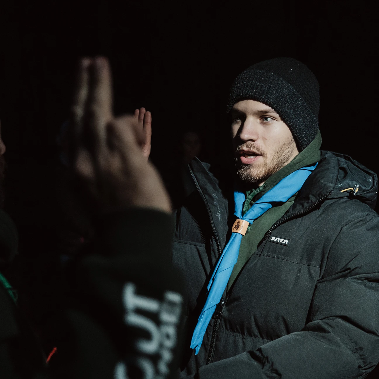 Un giovane indossa un cappello di lana e una giacca nera con una sciarpa azzurra annodata, mentre alza la mano destra come in un giuramento durante la notte.