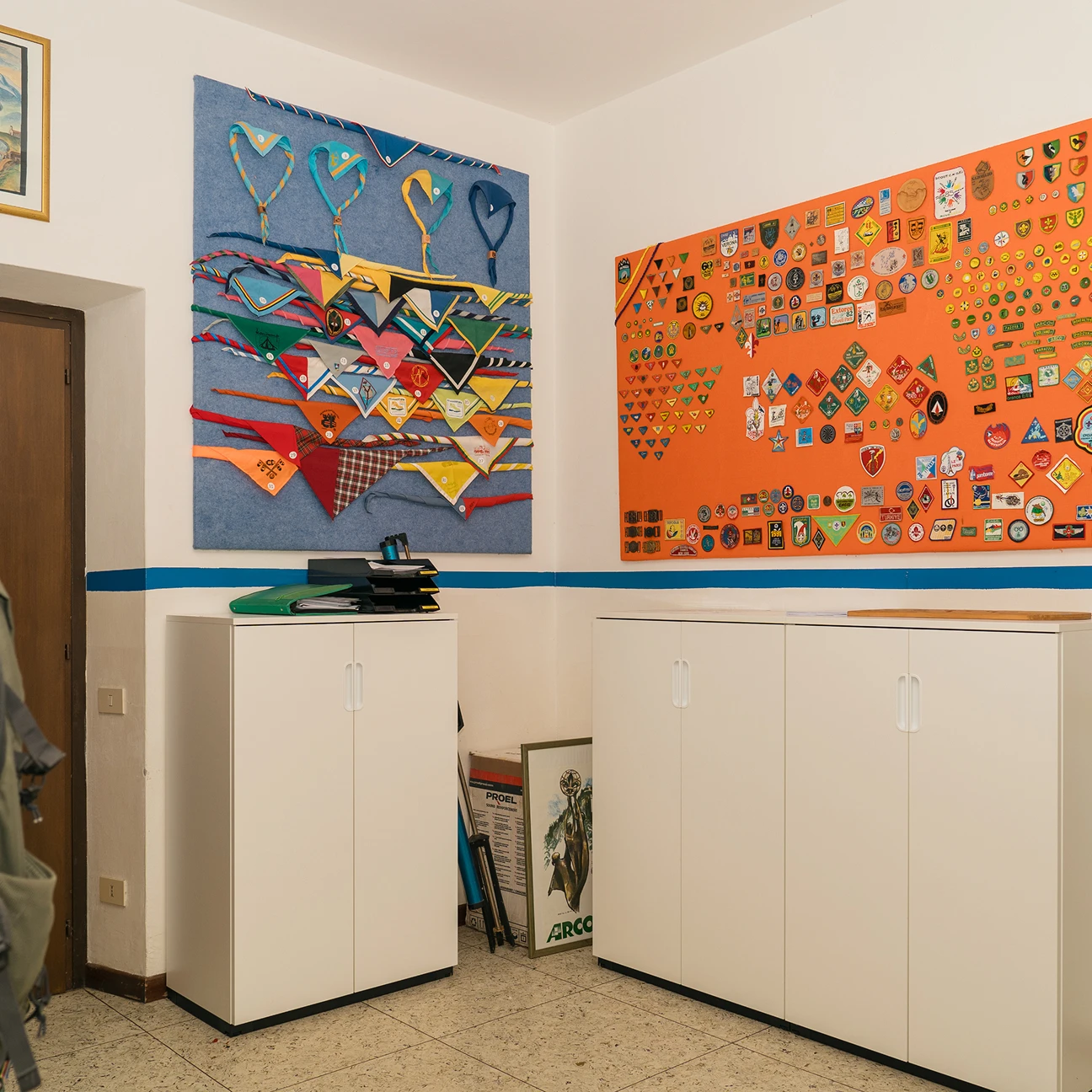 Parete di stanza con due pannelli appesi contenenti fazzoletti scout colorati e distintivi, armadietti bianchi in basso.