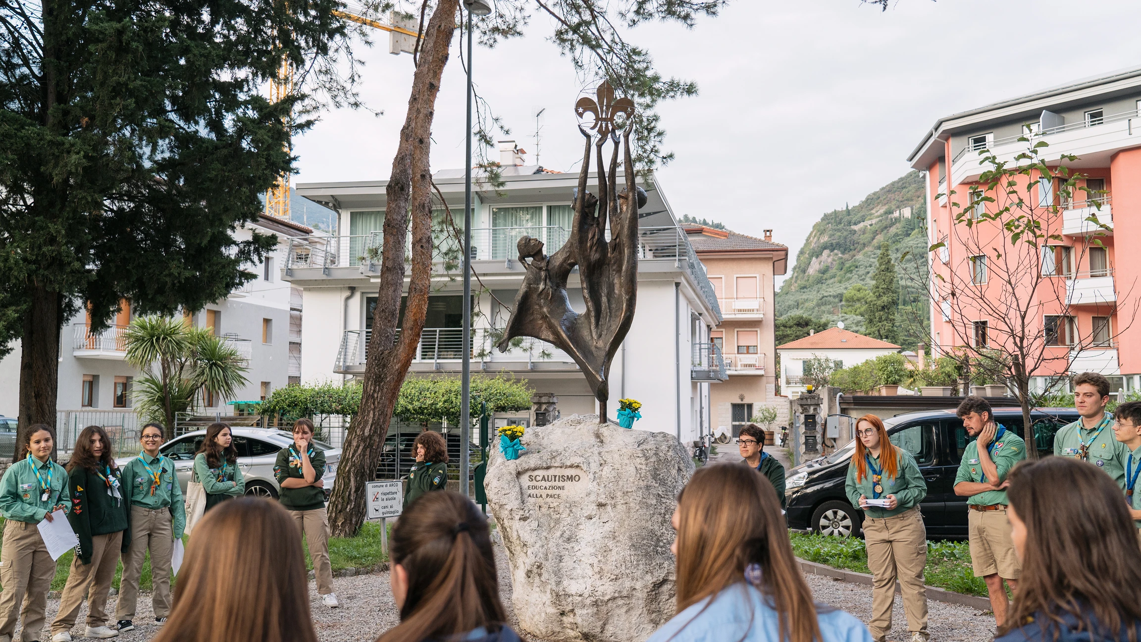 Un gruppo di scout in uniforme verde si trova attorno a una statua in bronzo su un masso con la scritta 'Scoutismo educazione alla pace' in un'area urbana con edifici e alberi.