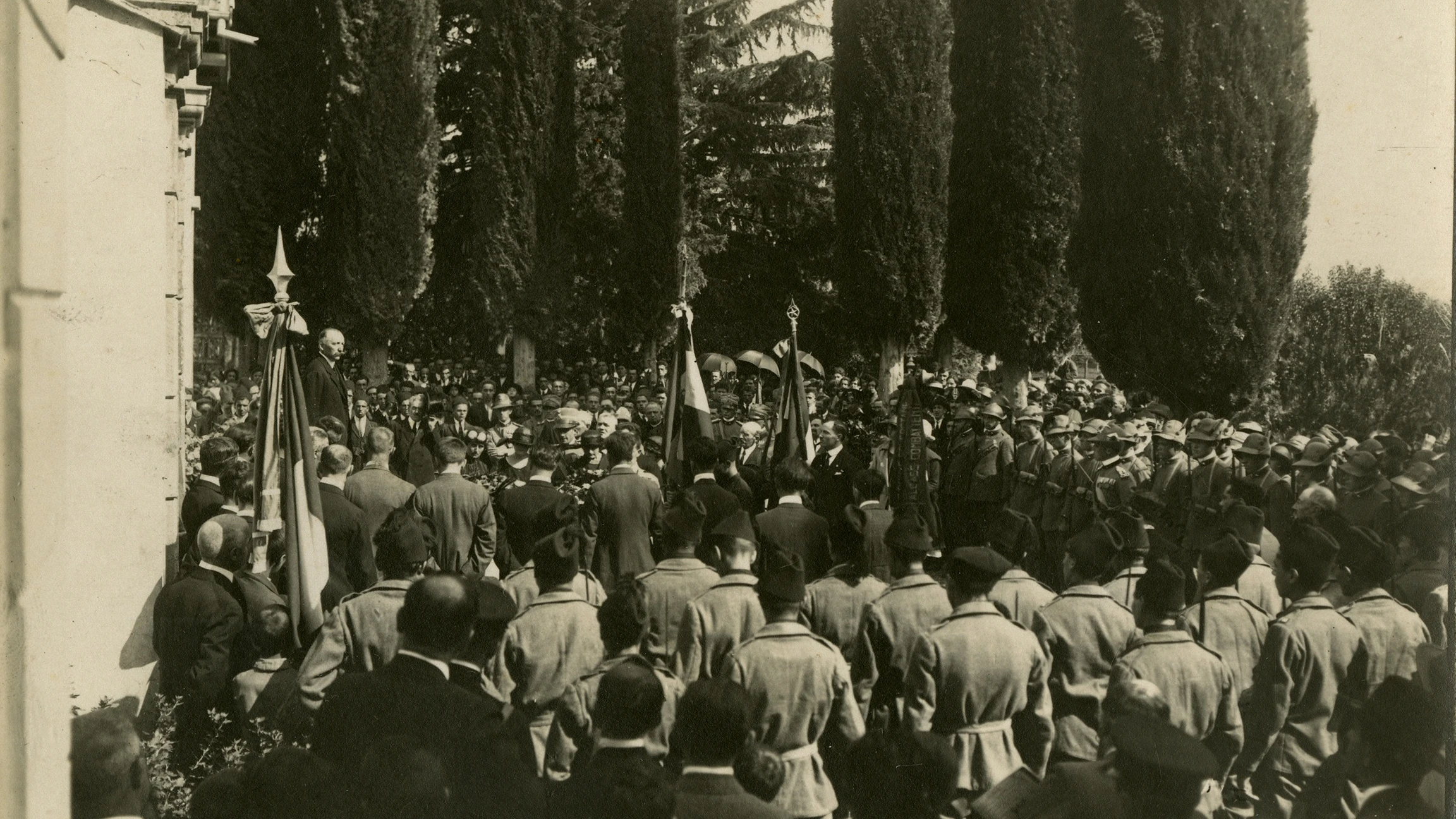 Foto storica in bianco e nero di una grande folla riunita all'aperto tra alberi alti, con molte persone in uniforme militare e altre in abiti formali che ascoltano un oratore su un podio.