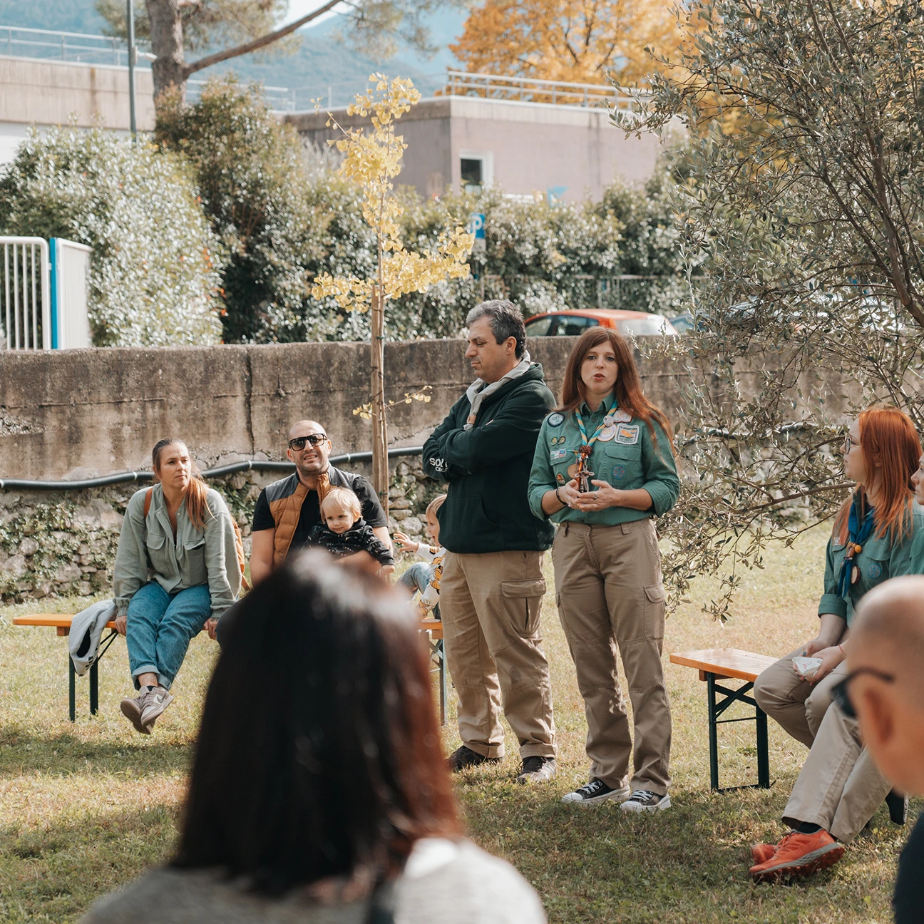 Una donna in uniforme da scout parla a un gruppo di persone all'aperto durante una giornata soleggiata.