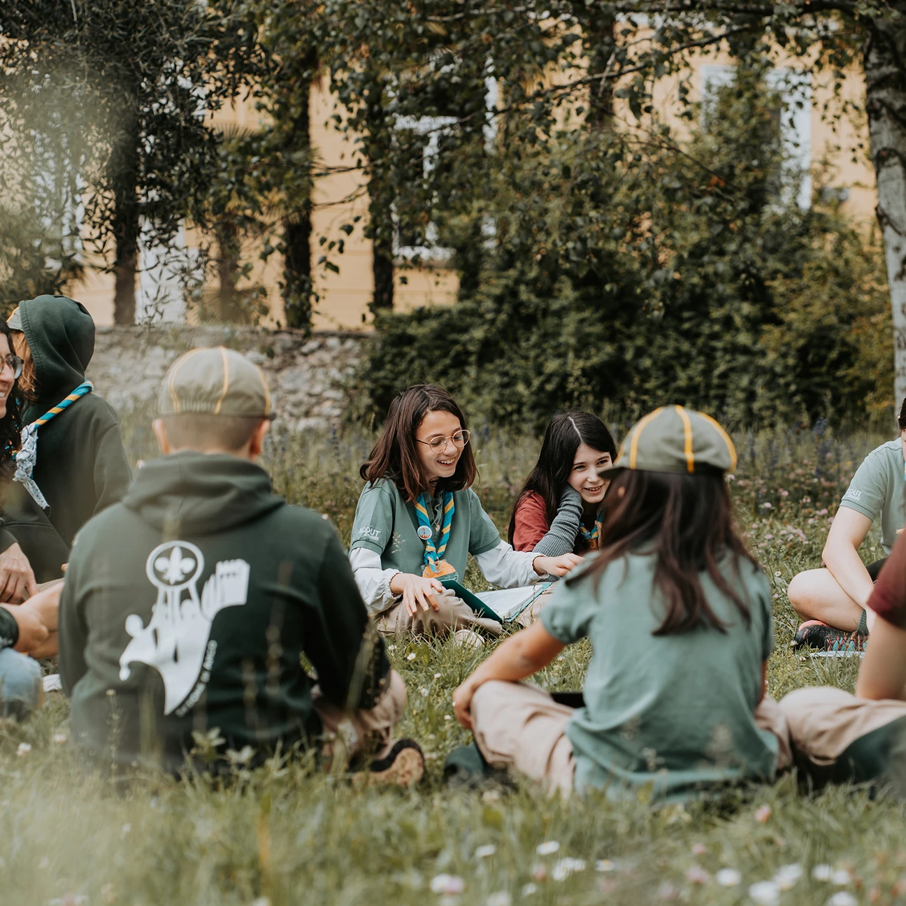 Bambini scout seduti in cerchio sull'erba in un parco, alcuni con sciarpe scout e cappellini, mentre ridono e parlano.