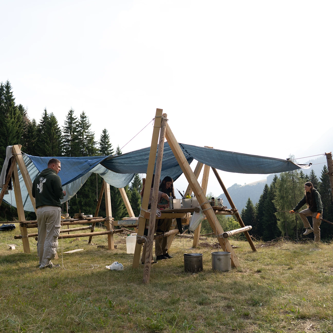Tre giovani allestiscono una cucina da campo con strutture di legno e teloni blu in un prato montano con alberi di conifere sullo sfondo.