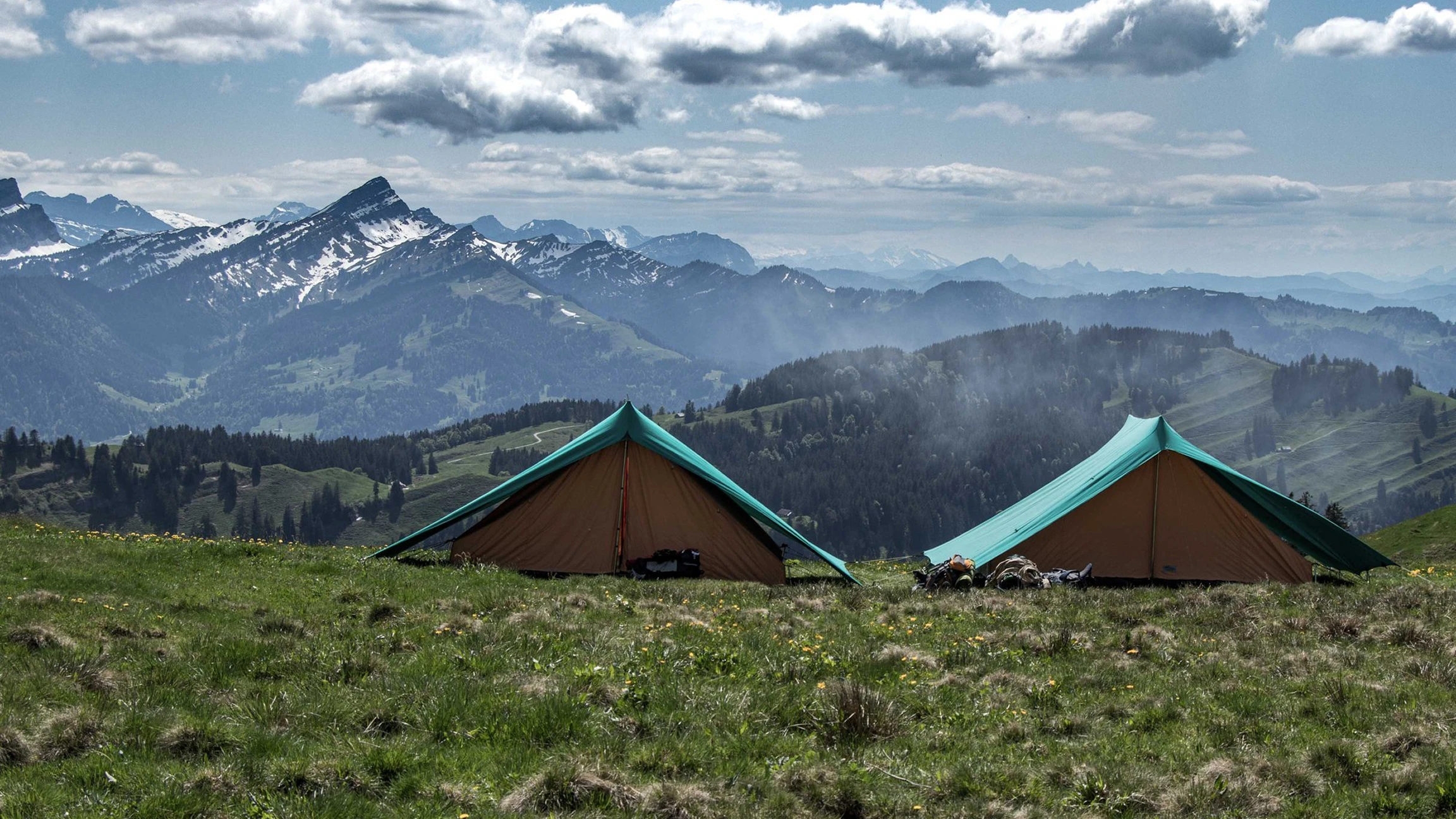 Due tende da campeggio su una radura erbosa con montagne innevate sullo sfondo sotto un cielo nuvoloso.