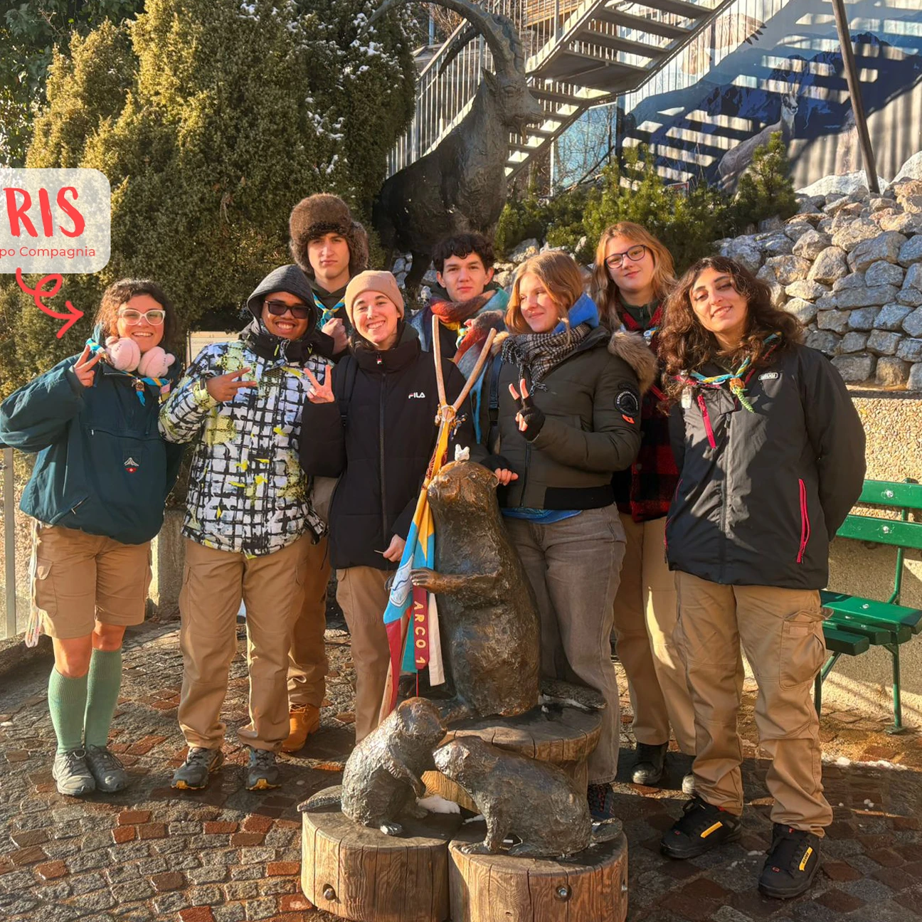 Un gruppo di giovani scout in abbigliamento invernale posano sorridenti intorno a una scultura di marmotte su pedane di legno all'aperto.