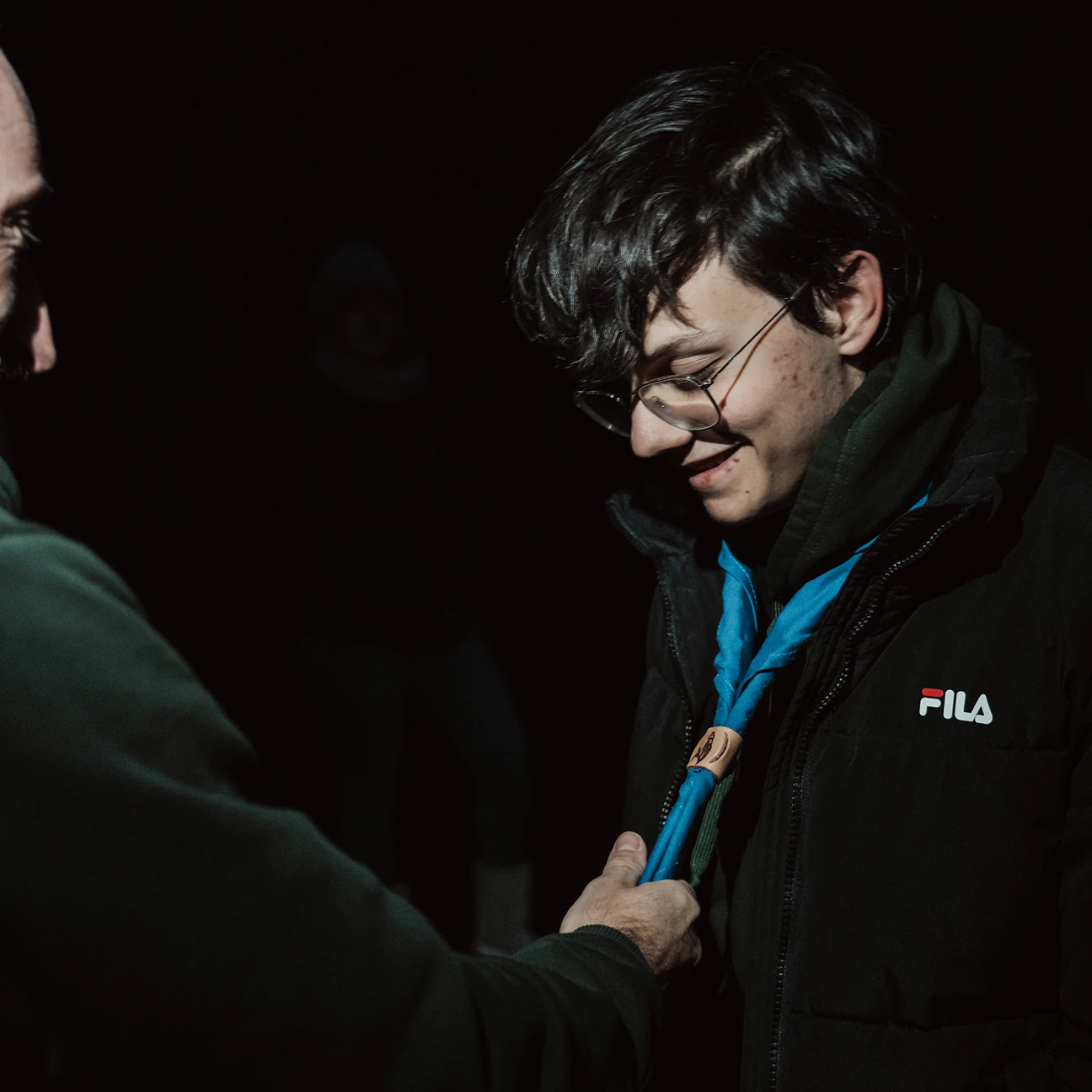 Un giovane con occhiali sorride mentre un'altra persona aggiusta la sua sciarpa azzurra da scout durante la notte.