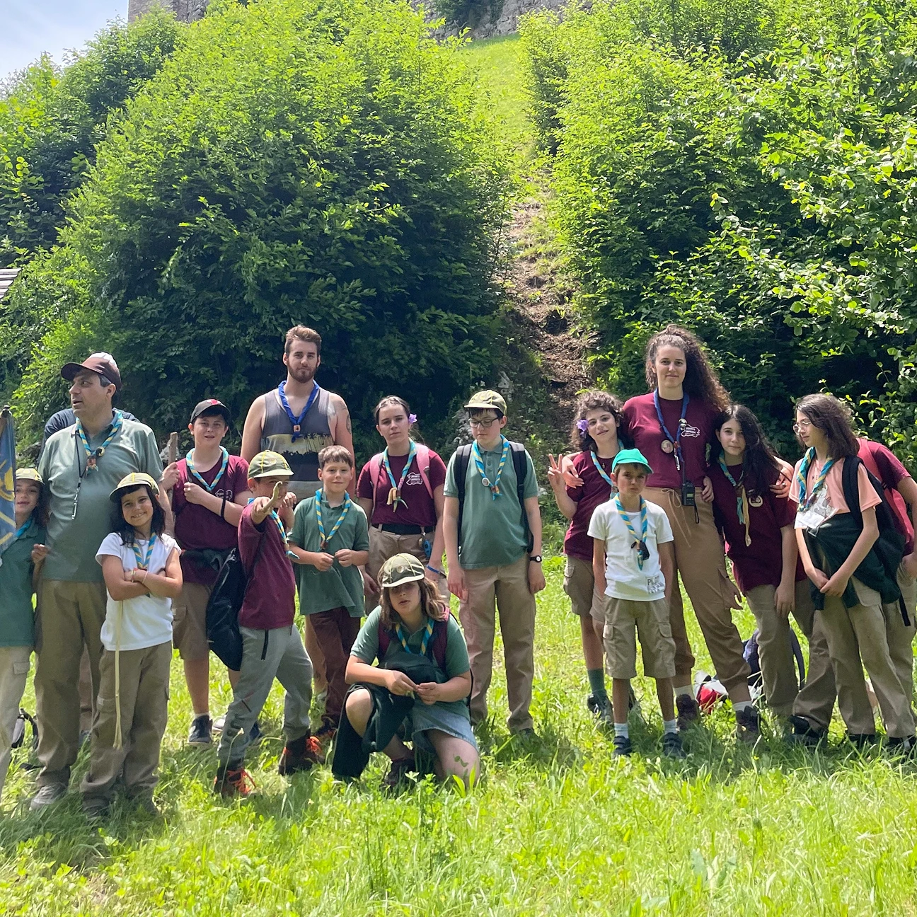Gruppo di scout giovani e adulti in uniformi colorate posano all'aperto su un prato verde con cespugli e alberi sullo sfondo.