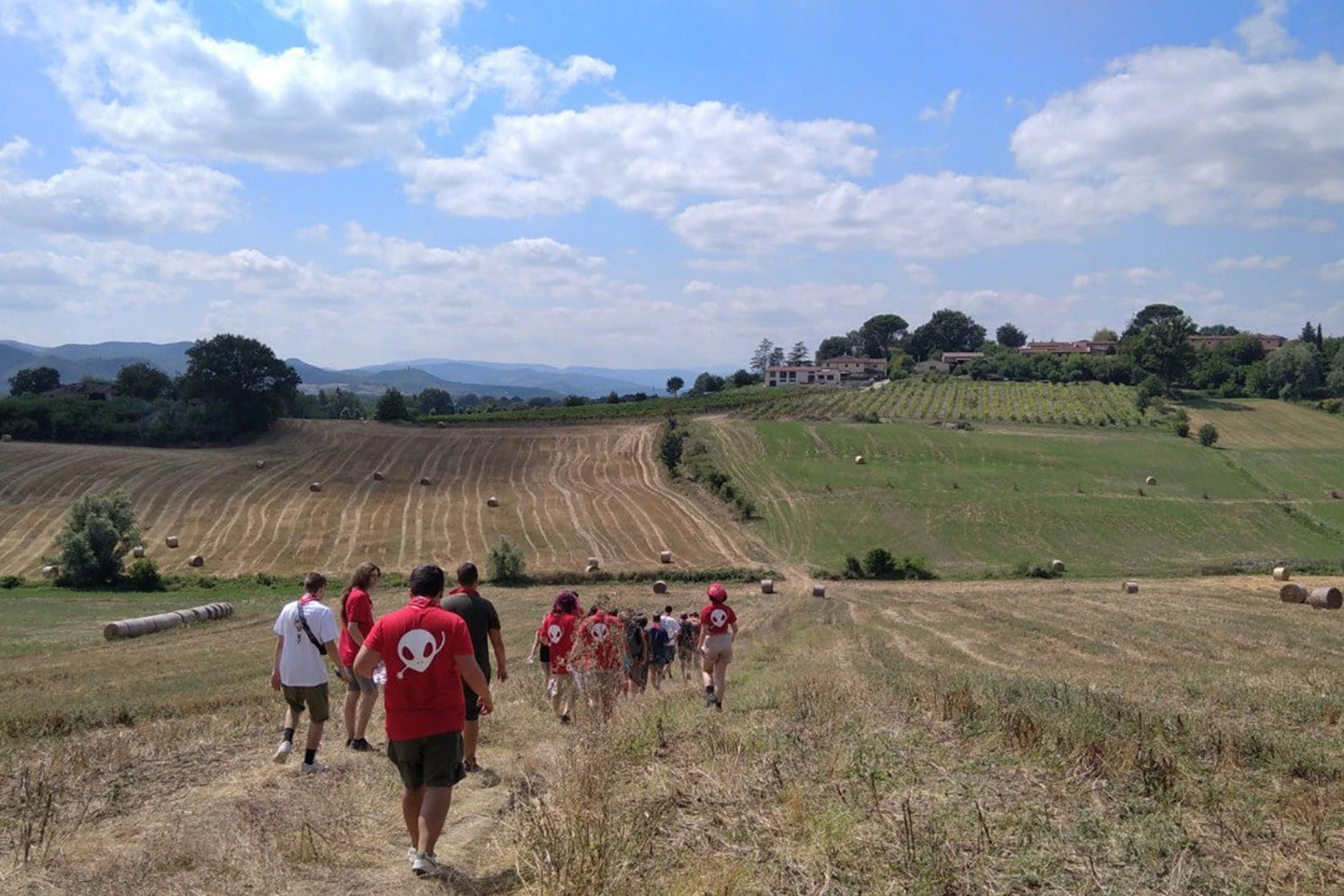 Un gruppo di persone cammina lungo un sentiero in una campagna con balle di fieno sparse e colline sullo sfondo.
