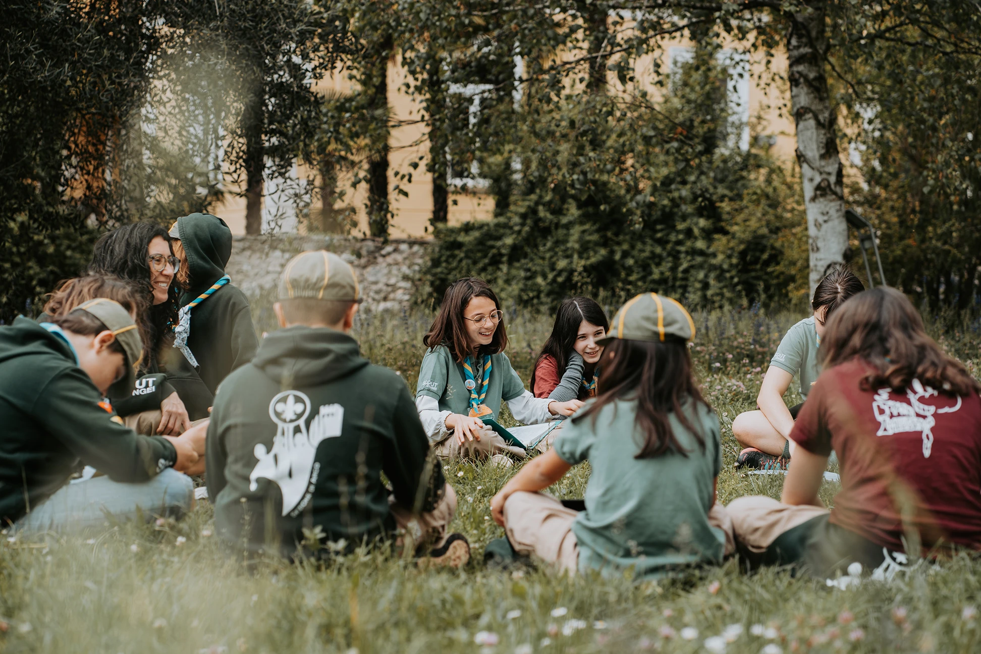 Un gruppo di ragazzi scout seduti in cerchio sull'erba in un parco mentre ridono e chiacchierano.