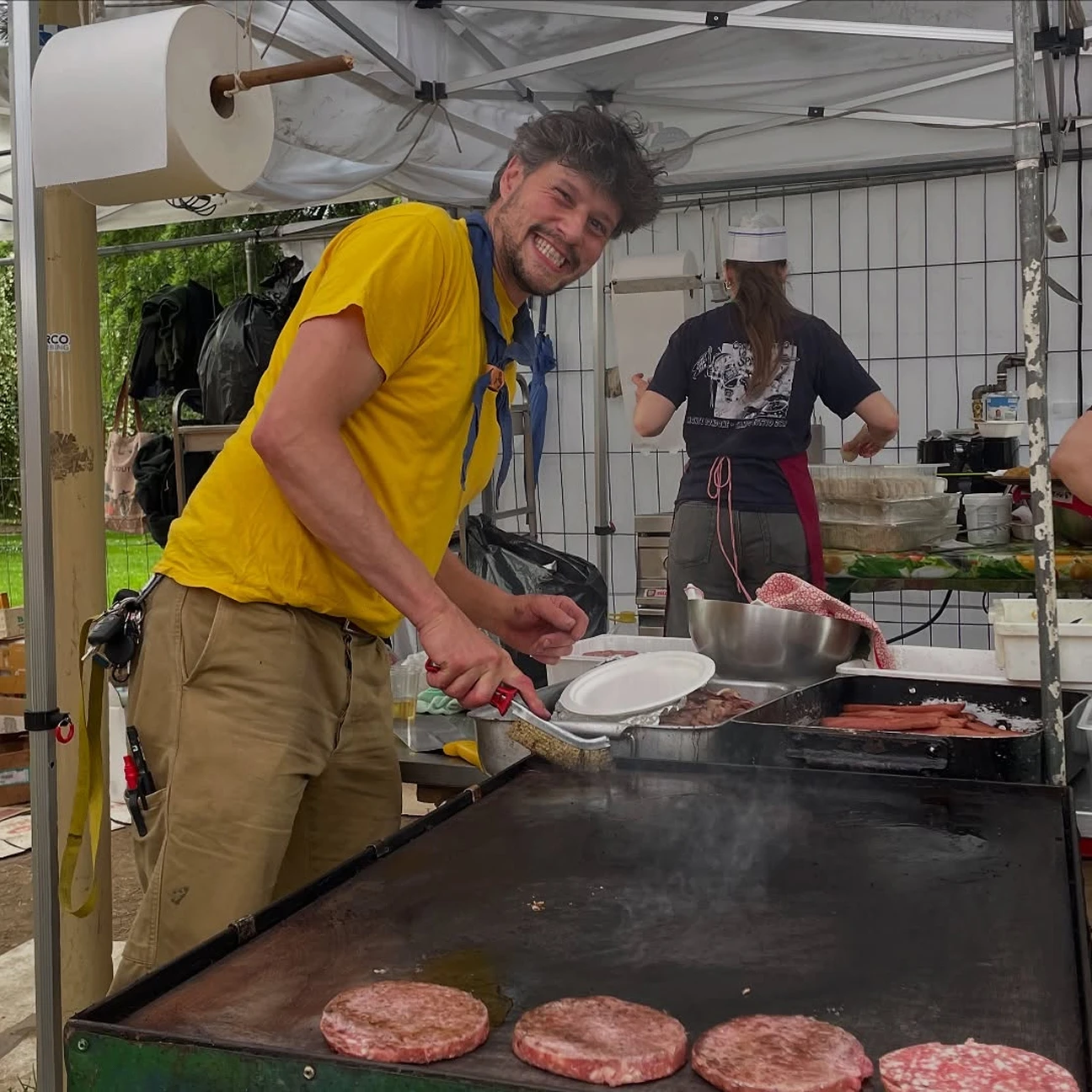 Uomo sorridente con maglietta gialla cucina hamburger su piastra in un chiosco all'aperto.