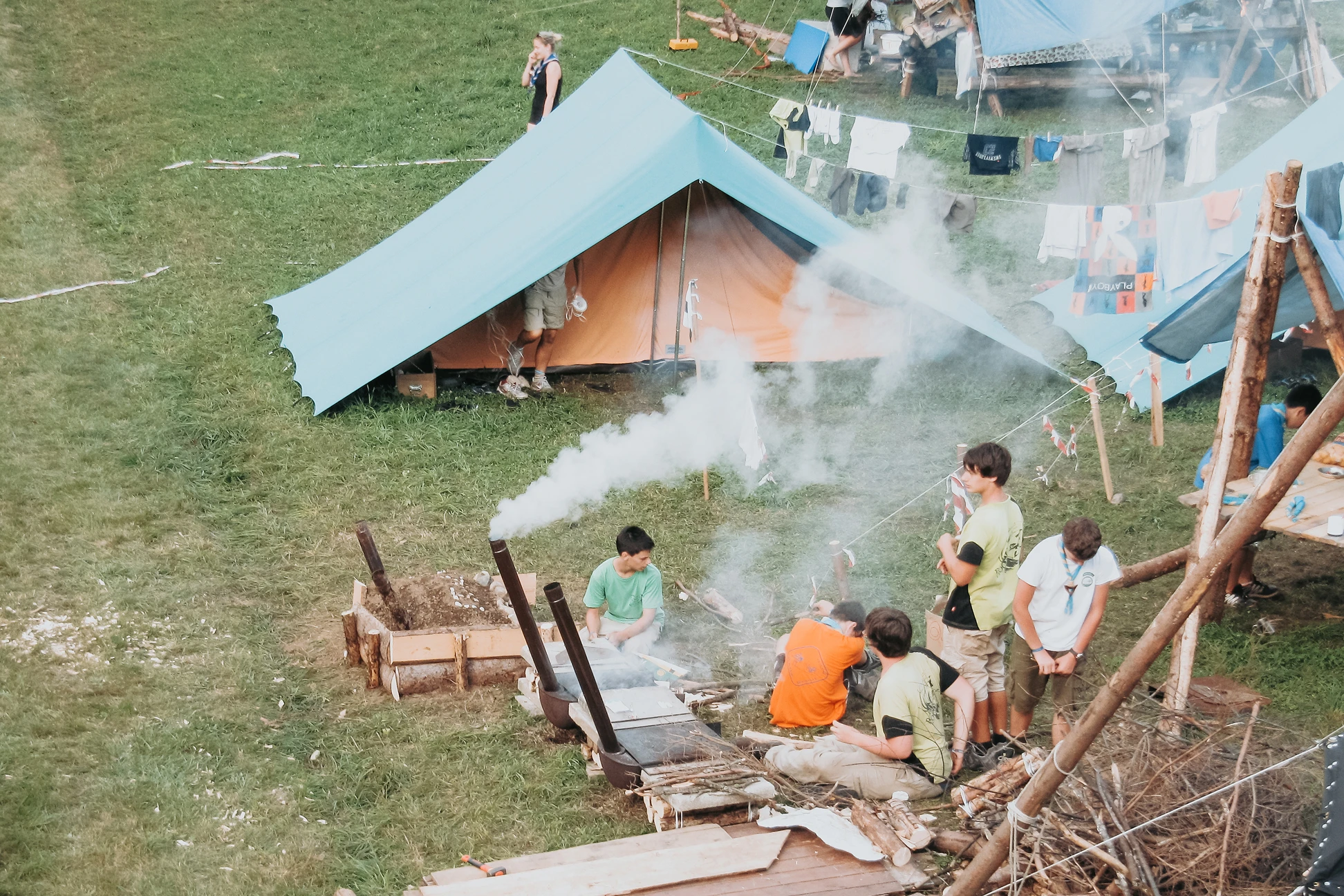 Ragazzi in campeggio accanto a tende blu con fumo che esce da una struttura fatta con legno e metallo, abiti stesi su una corda.