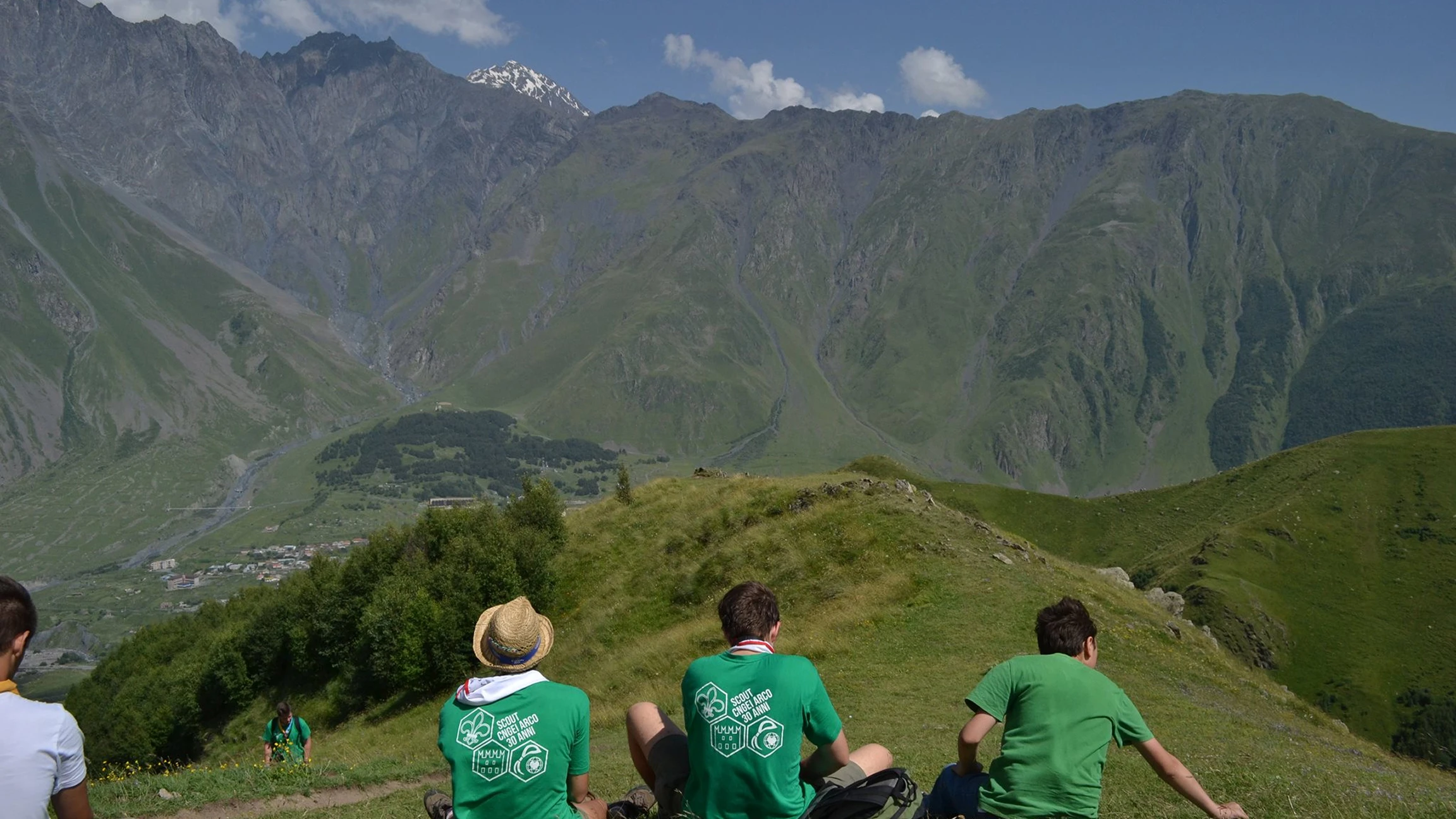 Tre ragazzi con magliette verdi seduti su una collina verde con una vista panoramica su montagne rocciose e un villaggio.