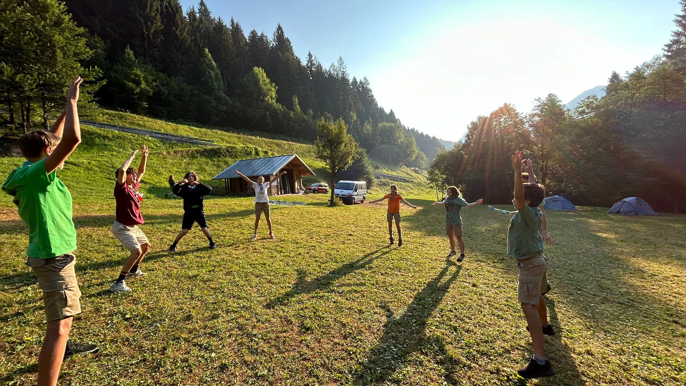 Un gruppo di bambini fa esercizi all'aperto su un prato verde con tende e un edificio di legno sullo sfondo sotto il sole.