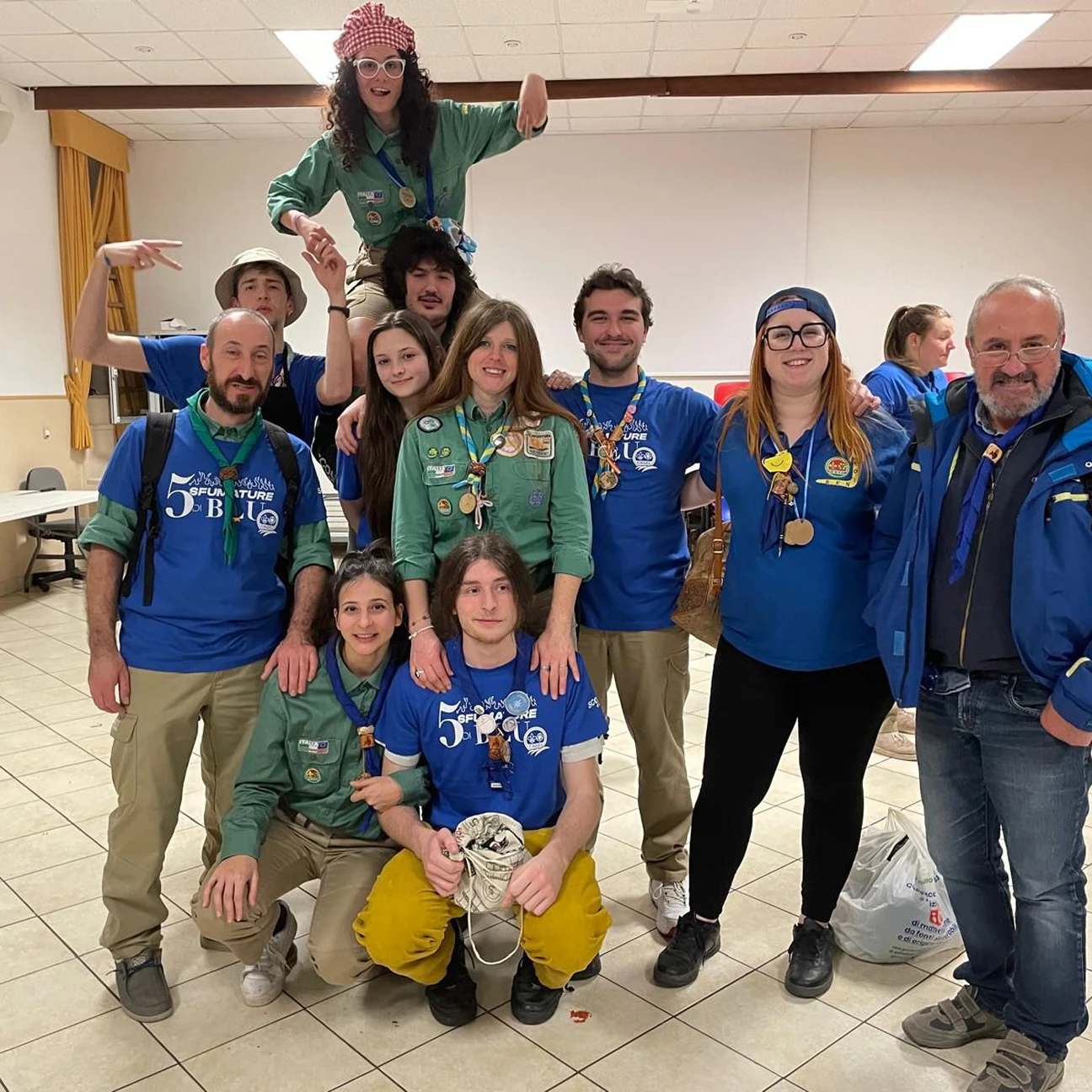Gruppo di giovani scout e adulti in uniforme con fazzoletti, alcuni sorridenti, in un ambiente interno.