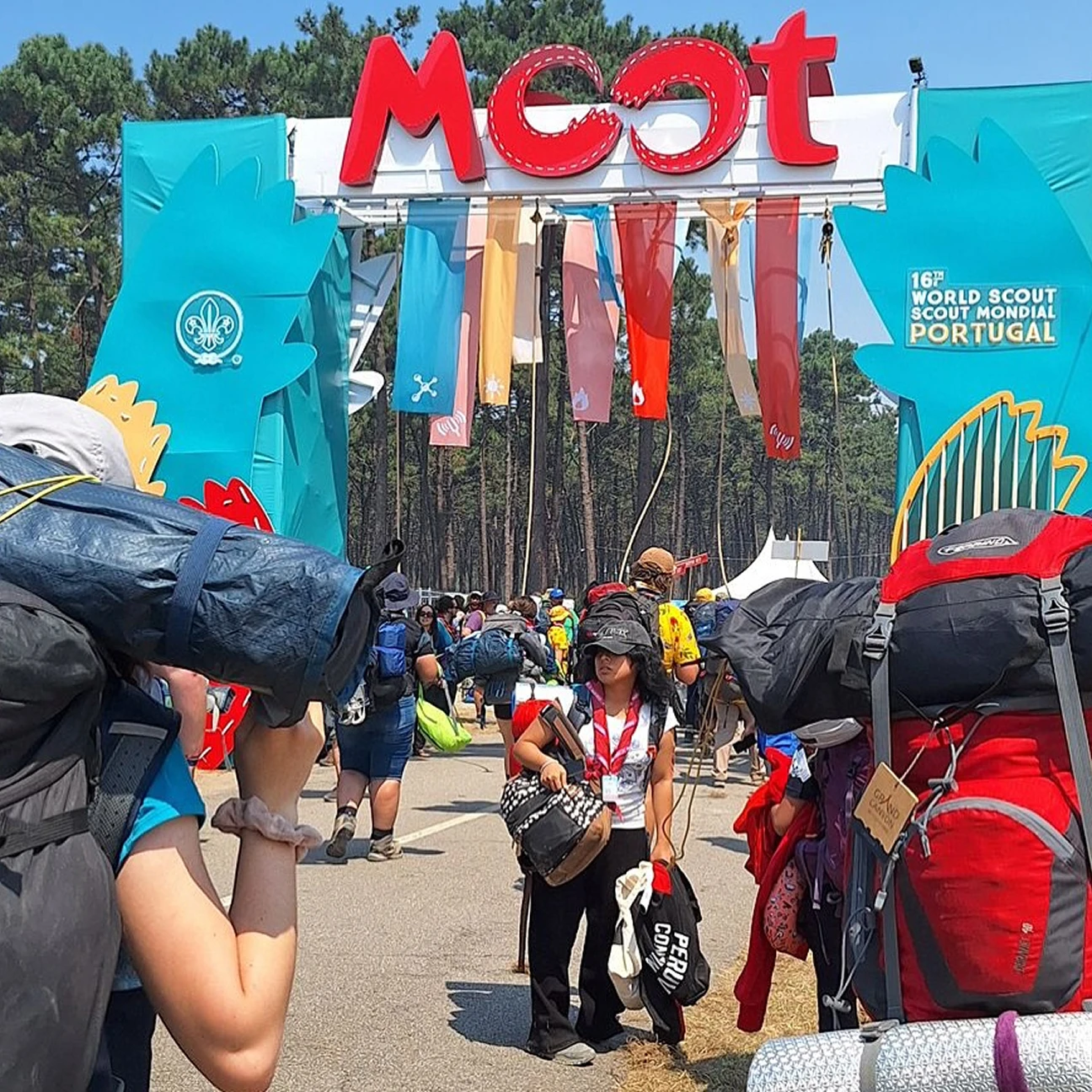Partecipanti Scout con zaini grandi sotto un arco colorato con scritta 'Moot' e bandiere, evento mondiale scout in Portogallo.