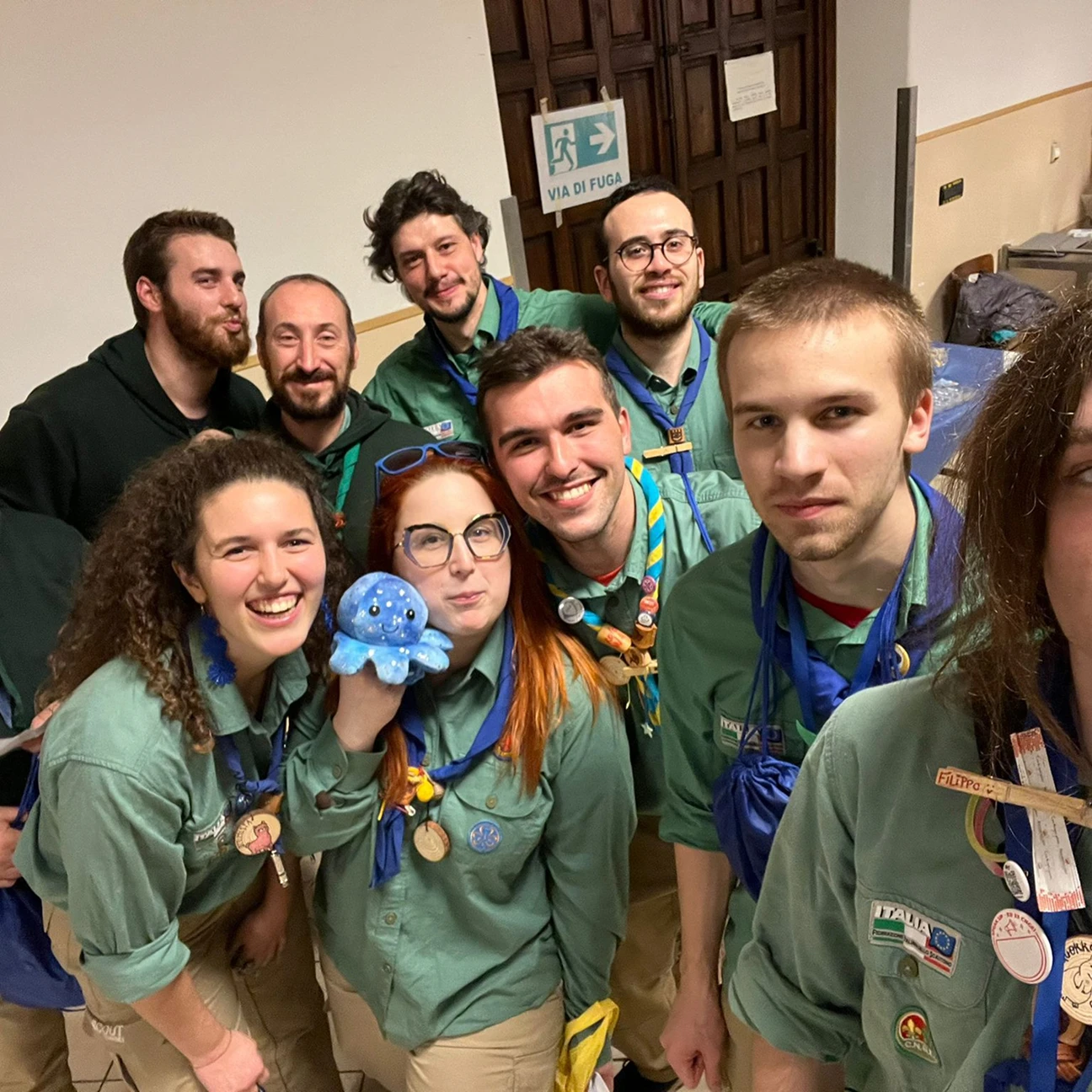 Gruppo di giovani scout con divise verdi e fazzoletti blu sorridenti durante una foto di gruppo al chiuso, una ragazza tiene un peluche polpo blu.