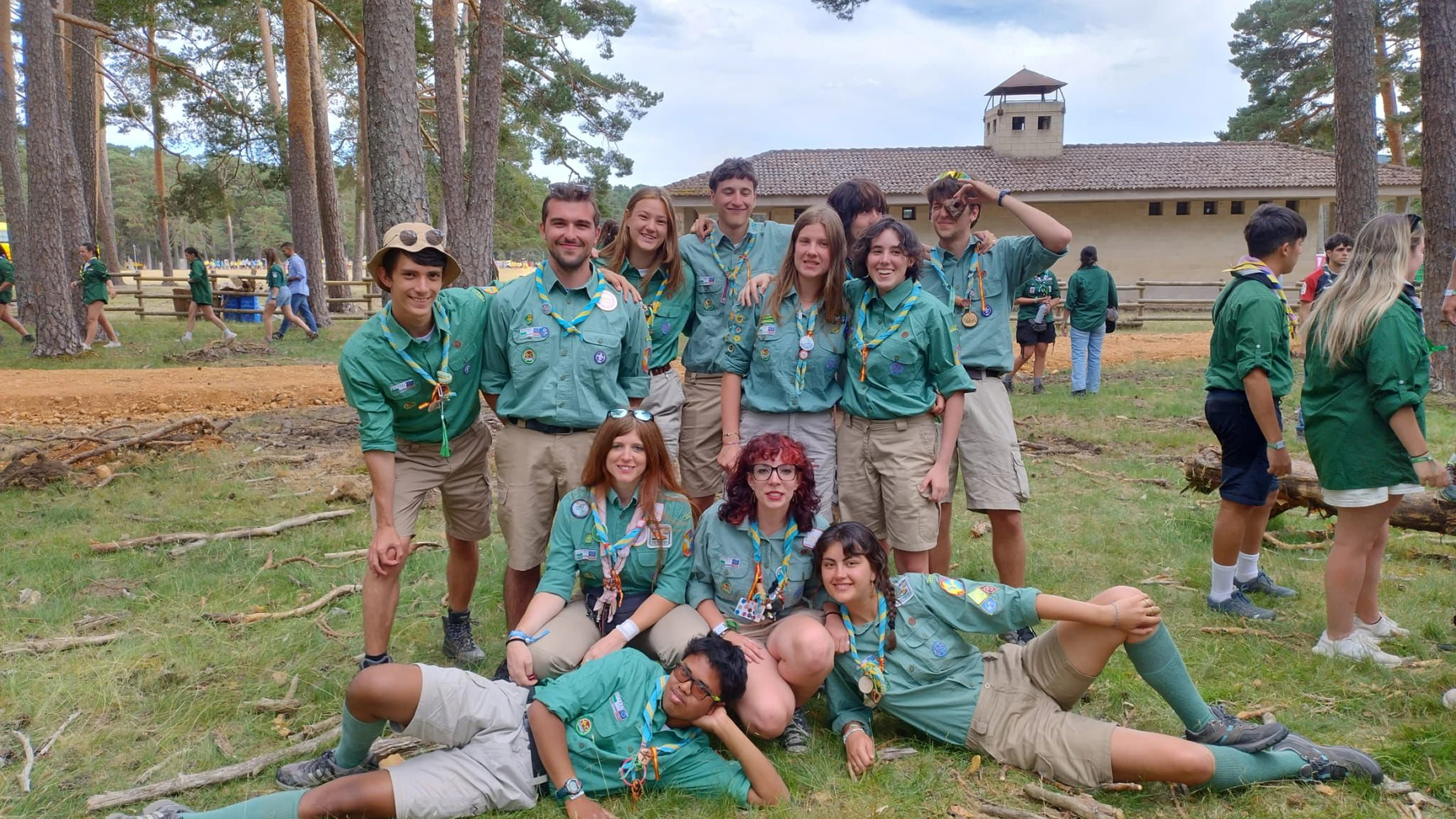 Gruppo di scout in uniforme verde e pantaloncini beige posa insieme su un prato in un ambiente boschivo con un edificio sullo sfondo.
