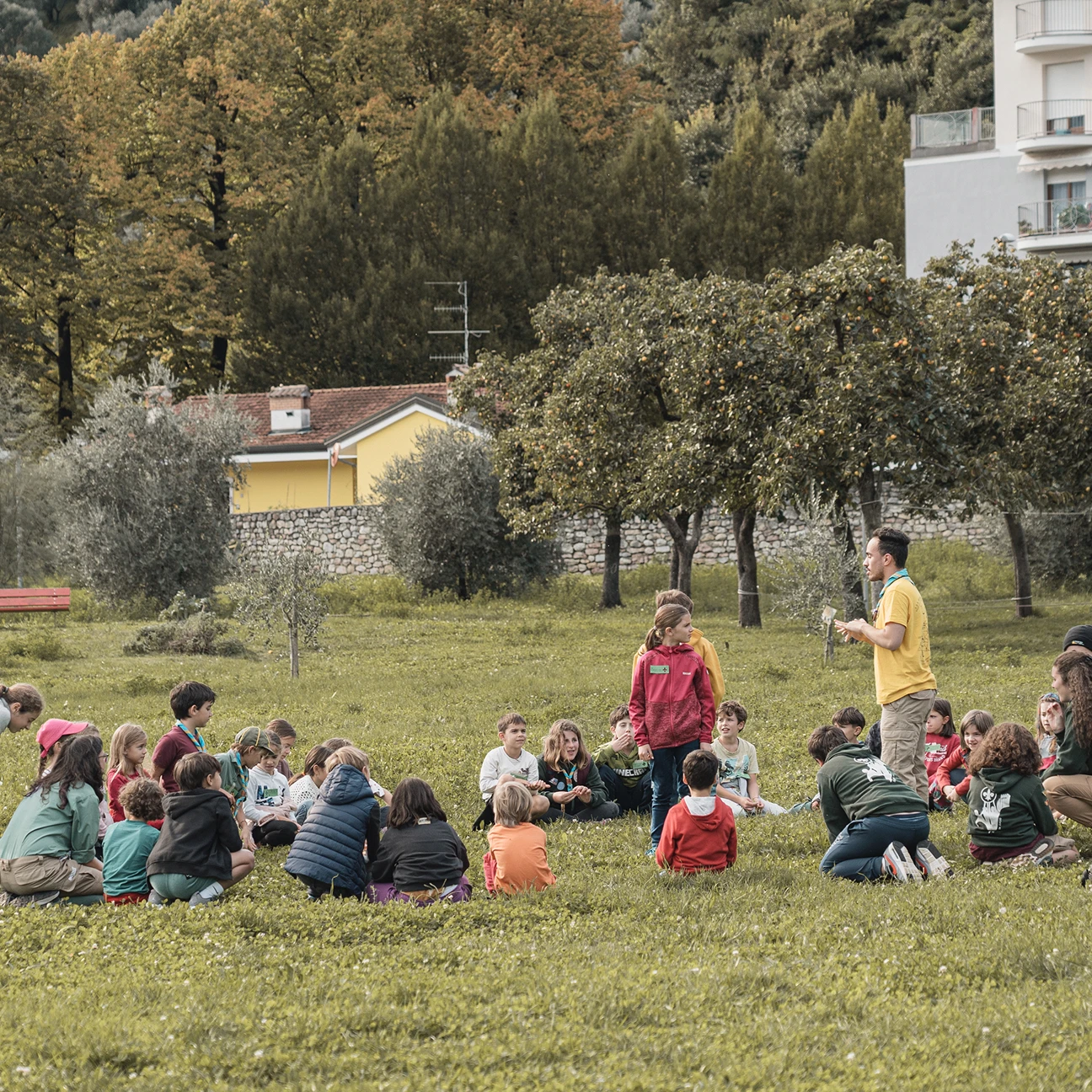 Un gruppo di bambini seduti sull'erba in cerchio durante un'attività all'aperto con un adulto che li guida, in un parco con alberi e case sullo sfondo.