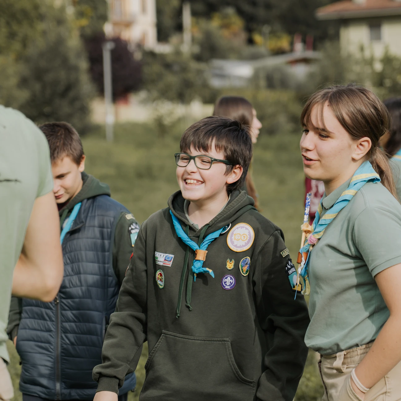 Ragazzo scout con occhiali e felpa verde scura con toppe sorridente all'aperto con altri scout.