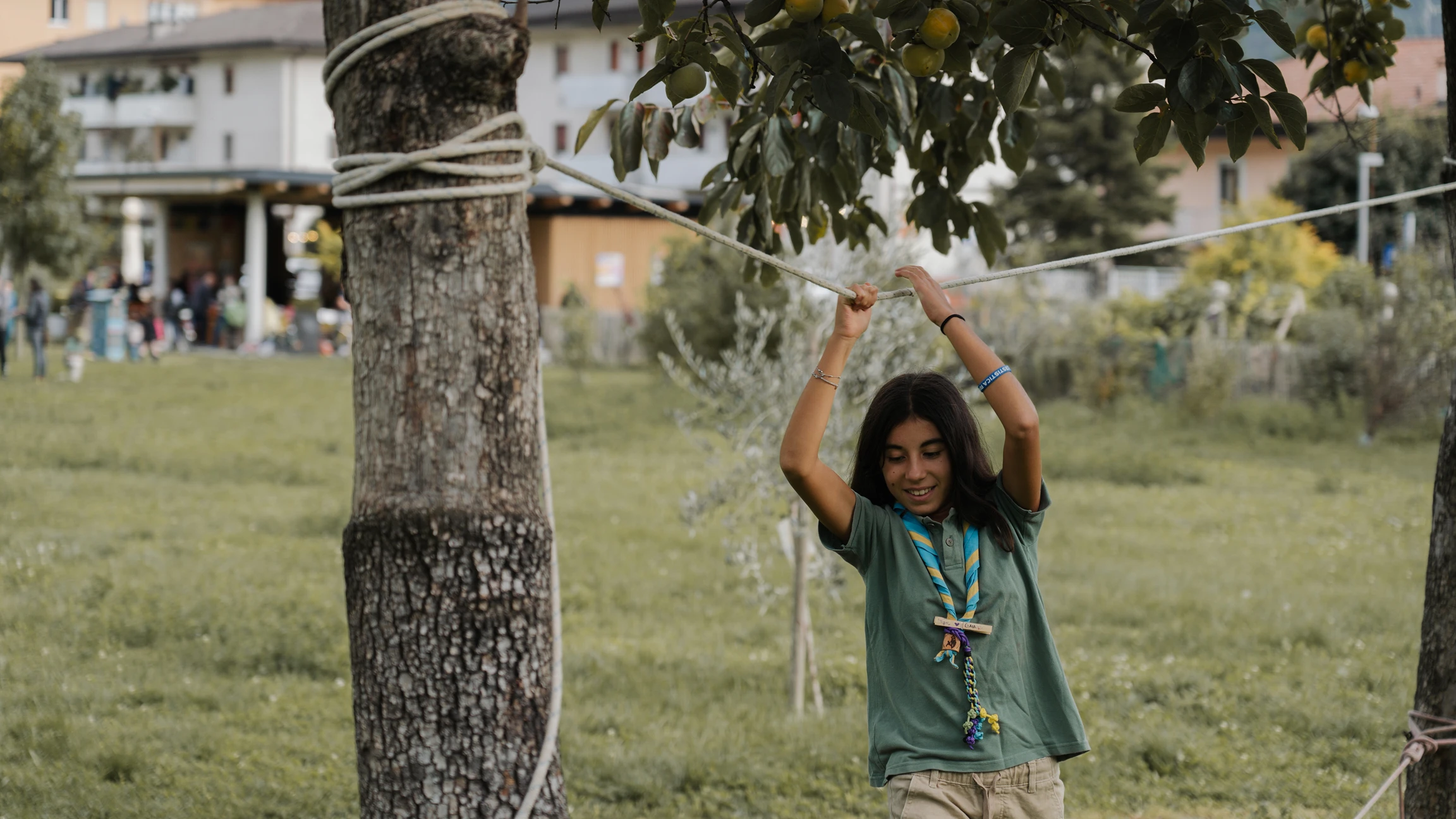 Ragazza scout con sciarpa blu e gialla tiene una corda fra due alberi in un prato verde.