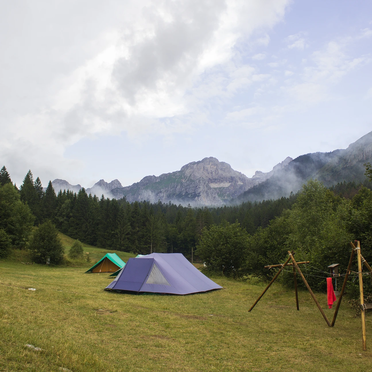 Tende da campeggio blu e verde in una radura erbosa con foresta e montagne nebbiose sullo sfondo.