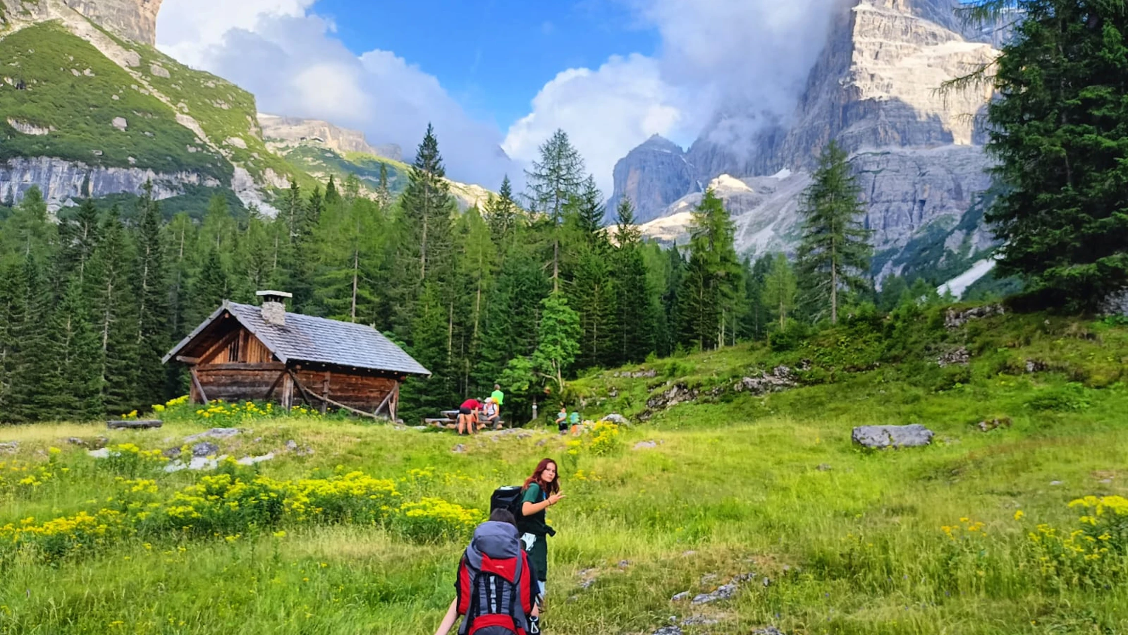 Escursionisti in una radura verde con fiori gialli, vicino a una baita di legno tra alberi e montagne rocciose con nuvole.