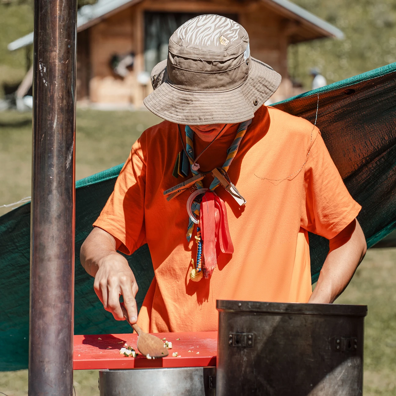 Ragazzo con cappello e maglietta arancione che taglia verdure su un tagliere rosso all'aperto.