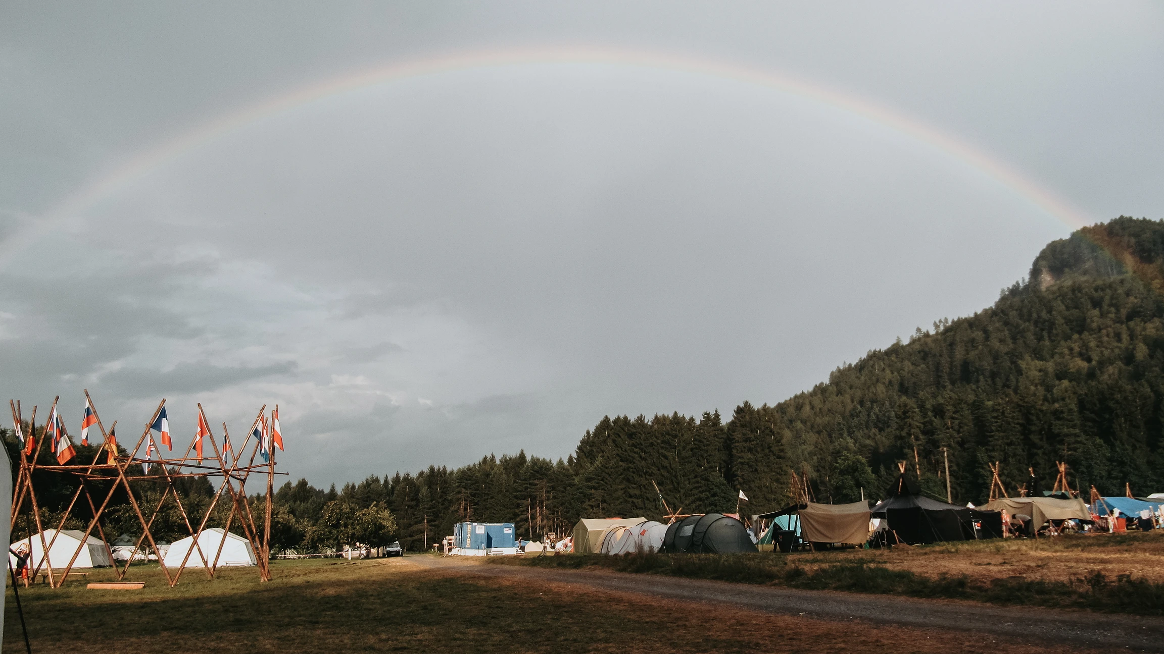 Campo scout con tende e una struttura in legno con bandiere, sotto un arcobaleno e cielo nuvoloso, con alberi e collina sullo sfondo.