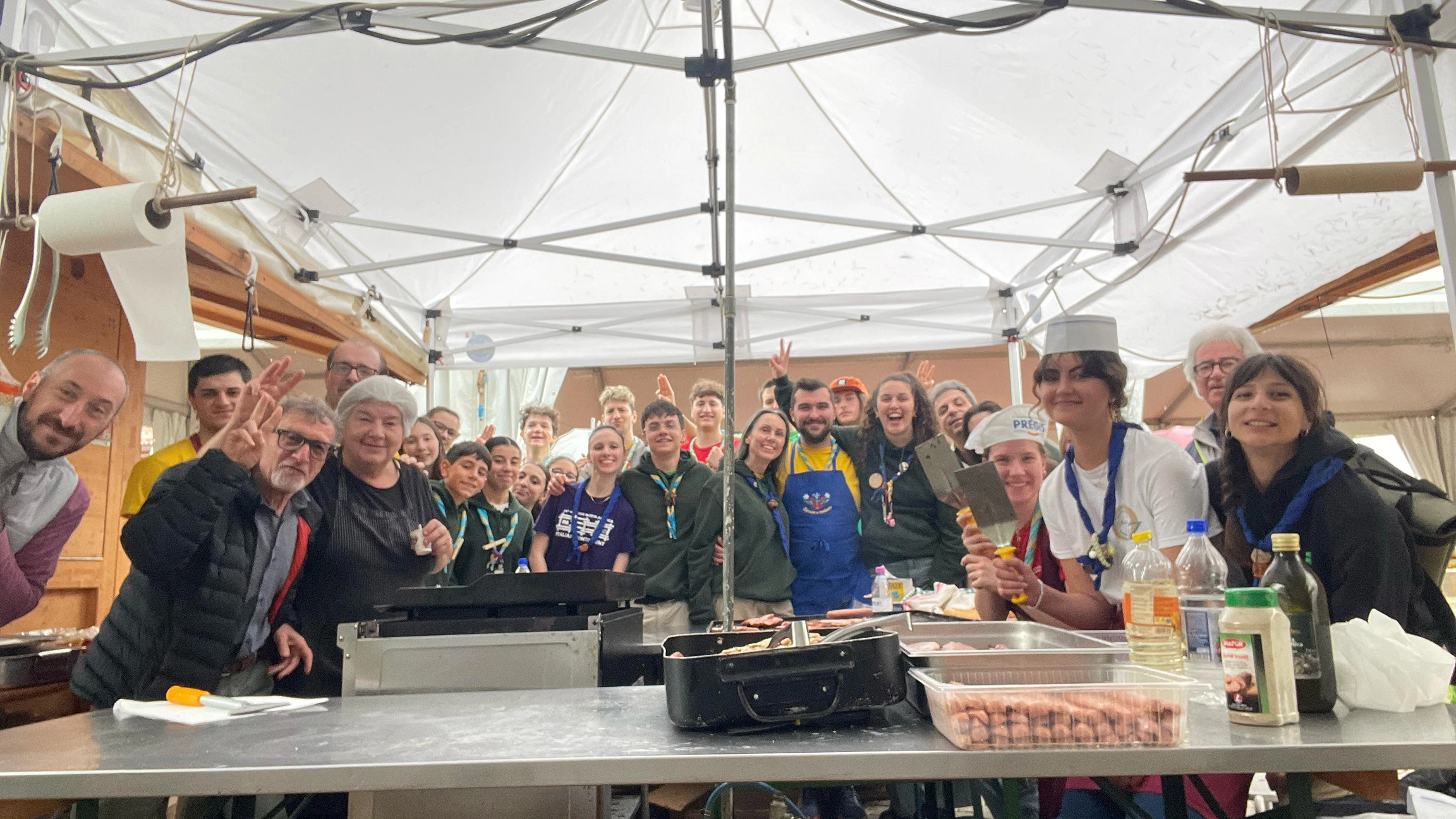 Un gruppo di persone sorridenti sotto una tenda, alcune con grembiuli e cappelli da cucina, pronte a cucinare insieme.