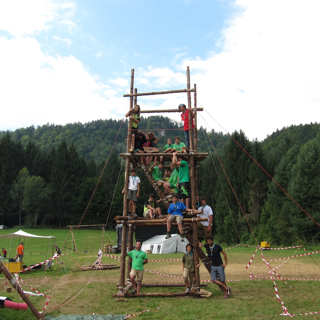 Gruppo di giovani seduti e in piedi su una struttura alta in legno all'aperto in un campo con alberi e colline sullo sfondo.