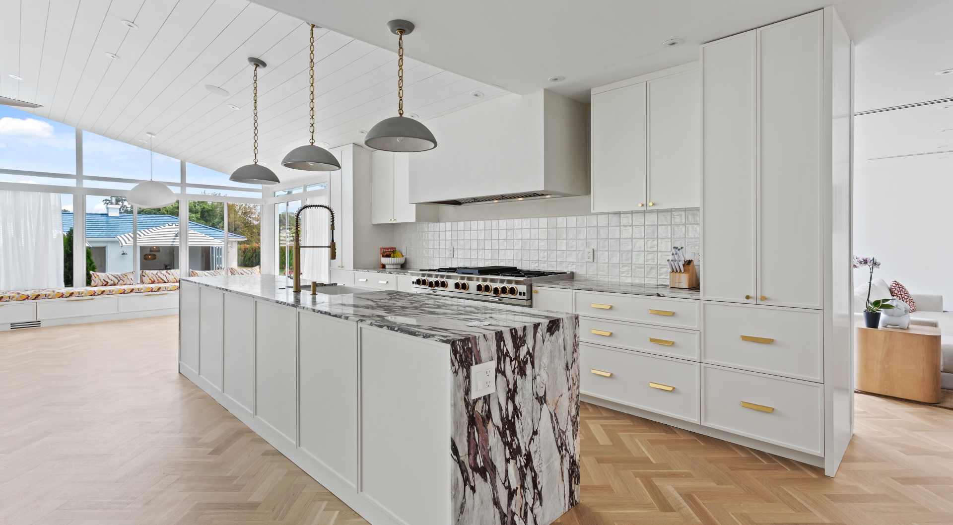 Bright modern kitchen with large marble island, white cabinetry with gold handles, and a seating area by floor-to-ceiling windows.