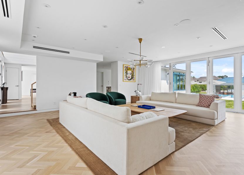Bright modern living room with two beige sofas, two green armchairs, wooden coffee table on brown rug, and large windows overlooking a backyard with a pool.