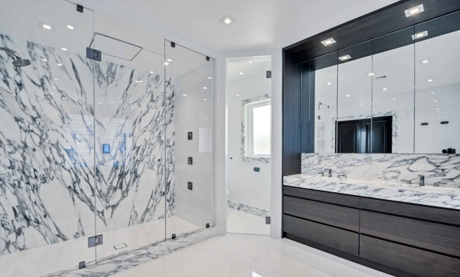 Modern bathroom with a large glass shower featuring white and gray marble walls, a double sink vanity with black cabinetry, and a large mirror.